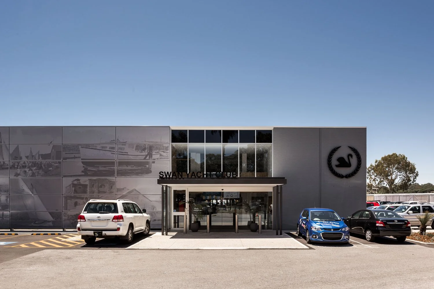 Front exterior of Swan Yacht Club building with cars parked in front, including white, blue, and black cars, under a clear blue sky.