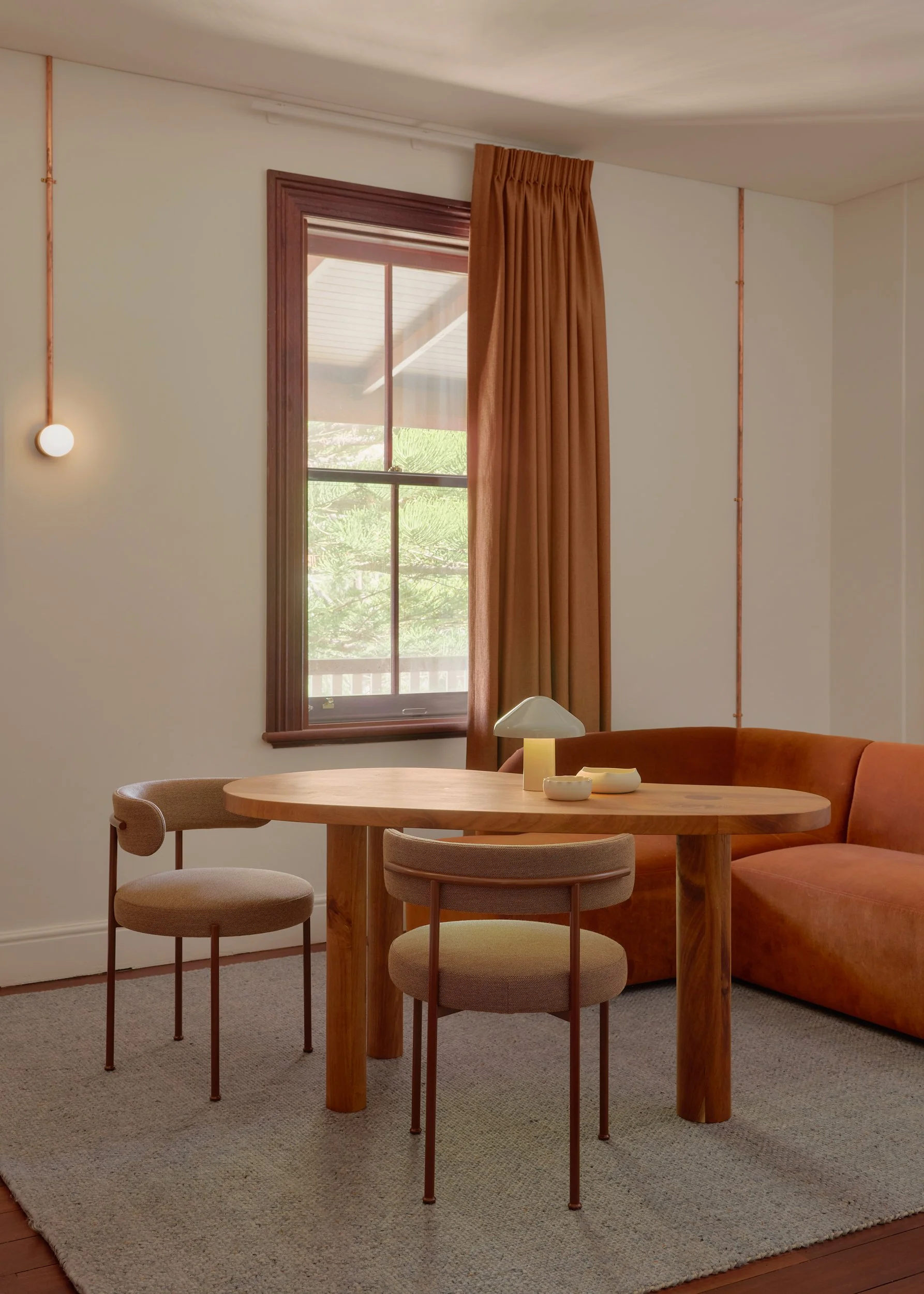 A cozy living room with a round wooden dining table, two beige upholstered chairs, a burnt orange sofa, a window with brown curtains, and a small modern lamp on the table.