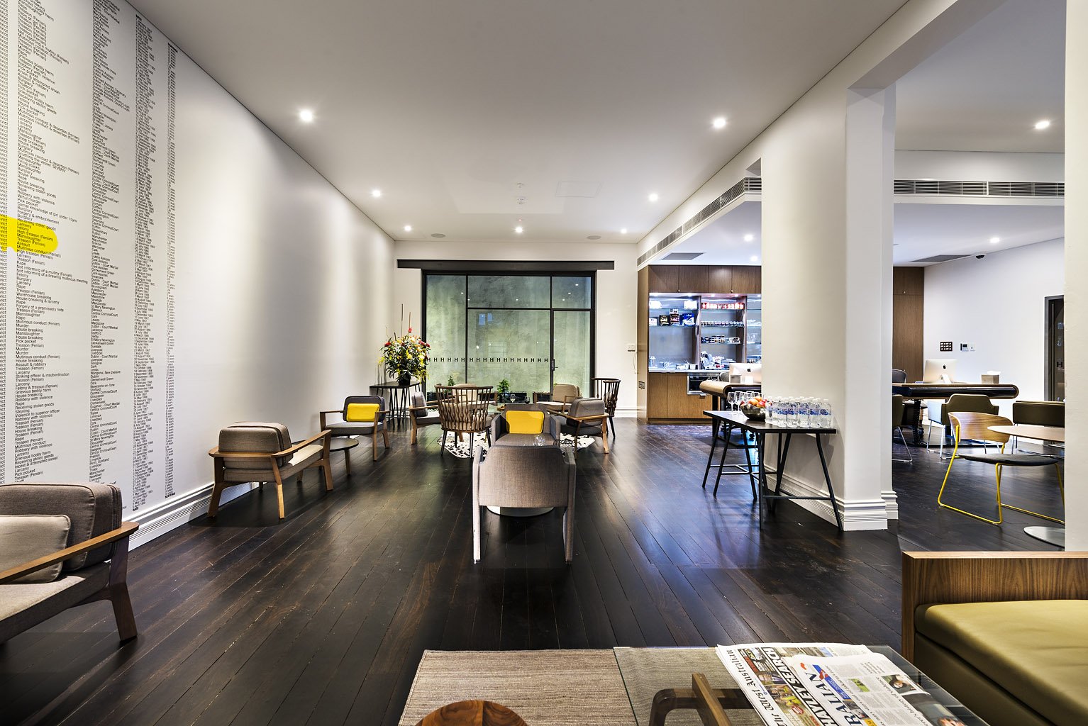 Modern cafe with dark hardwood floors, white walls, and various seating options including armchairs, benches, and small tables. A large window at the back provides natural light, and a beverage station with bottled water is on the right. The space is