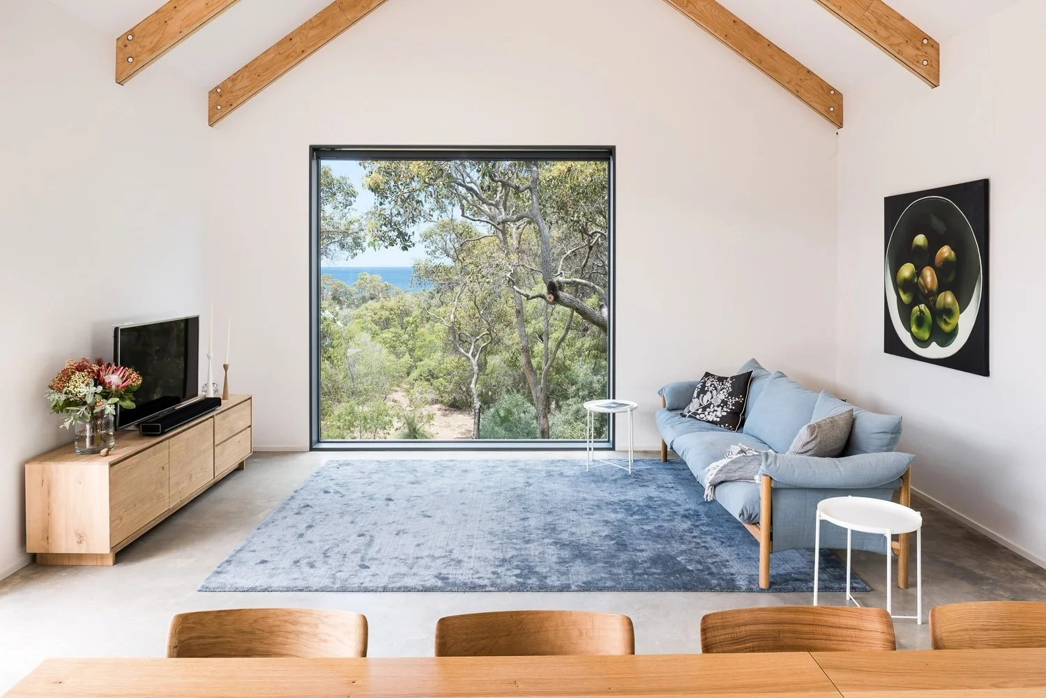 Interior of a minimalist living room with a large window showing trees and a blue sky outside, a light blue sofa with pillows, a wooden TV stand with a TV and flowers, and a gray rug.