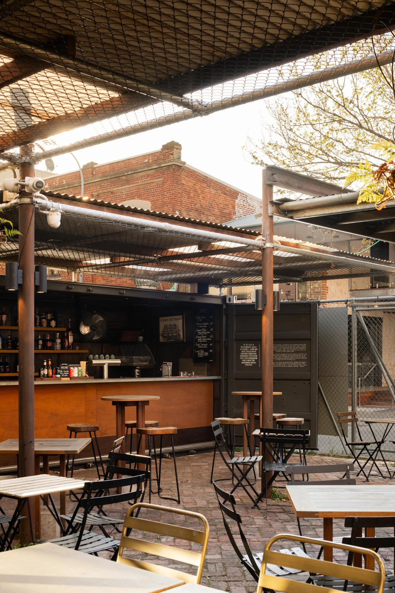 An outdoor bar area with wooden tables and chairs, a brick building in the background, and a covered patio with metal poles and netting overhead.