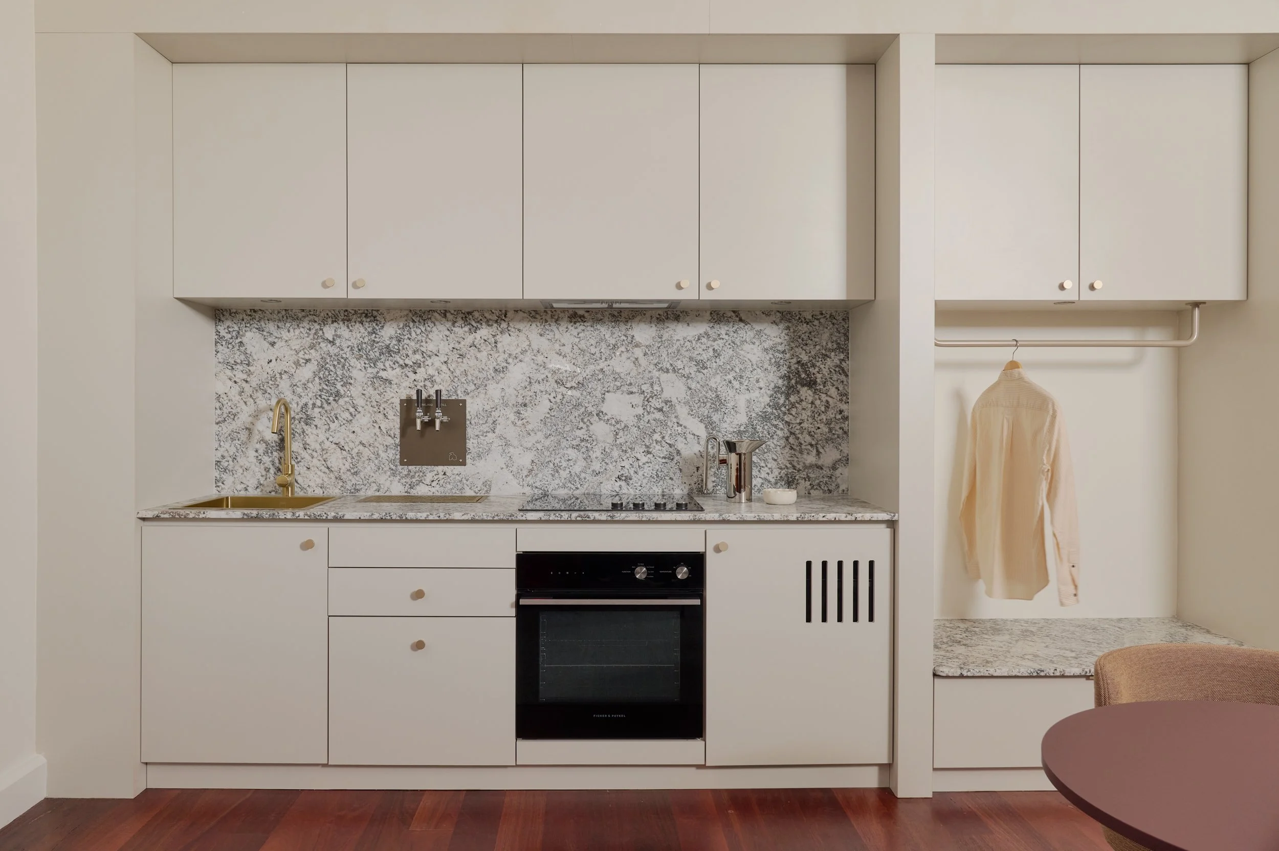 Minimalist kitchen with white cabinets, marble backsplash, and a black oven, adjacent to a small nook with a hanging shirt and marble bench.