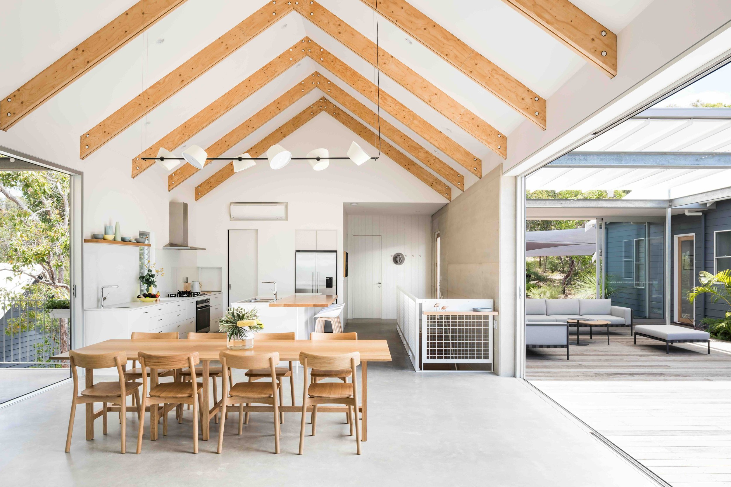 Open concept living space with kitchen, dining area, and outdoor patio. The high ceiling has exposed wooden beams, and large sliding glass doors open to a wooden deck with outdoor seating.