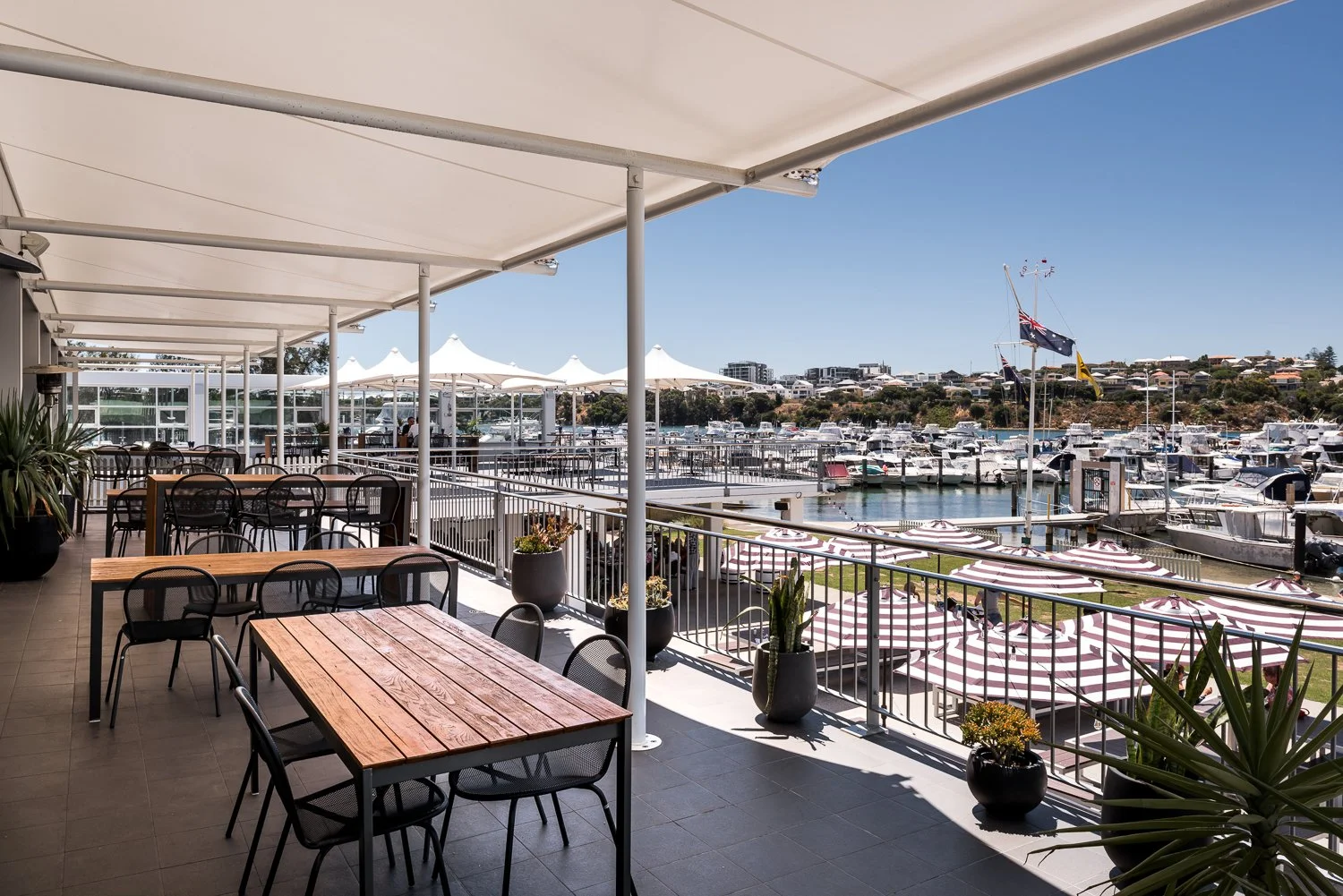 An outdoor restaurant or cafe patio overlooking a marina filled with boats, with striped umbrellas and potted plants.