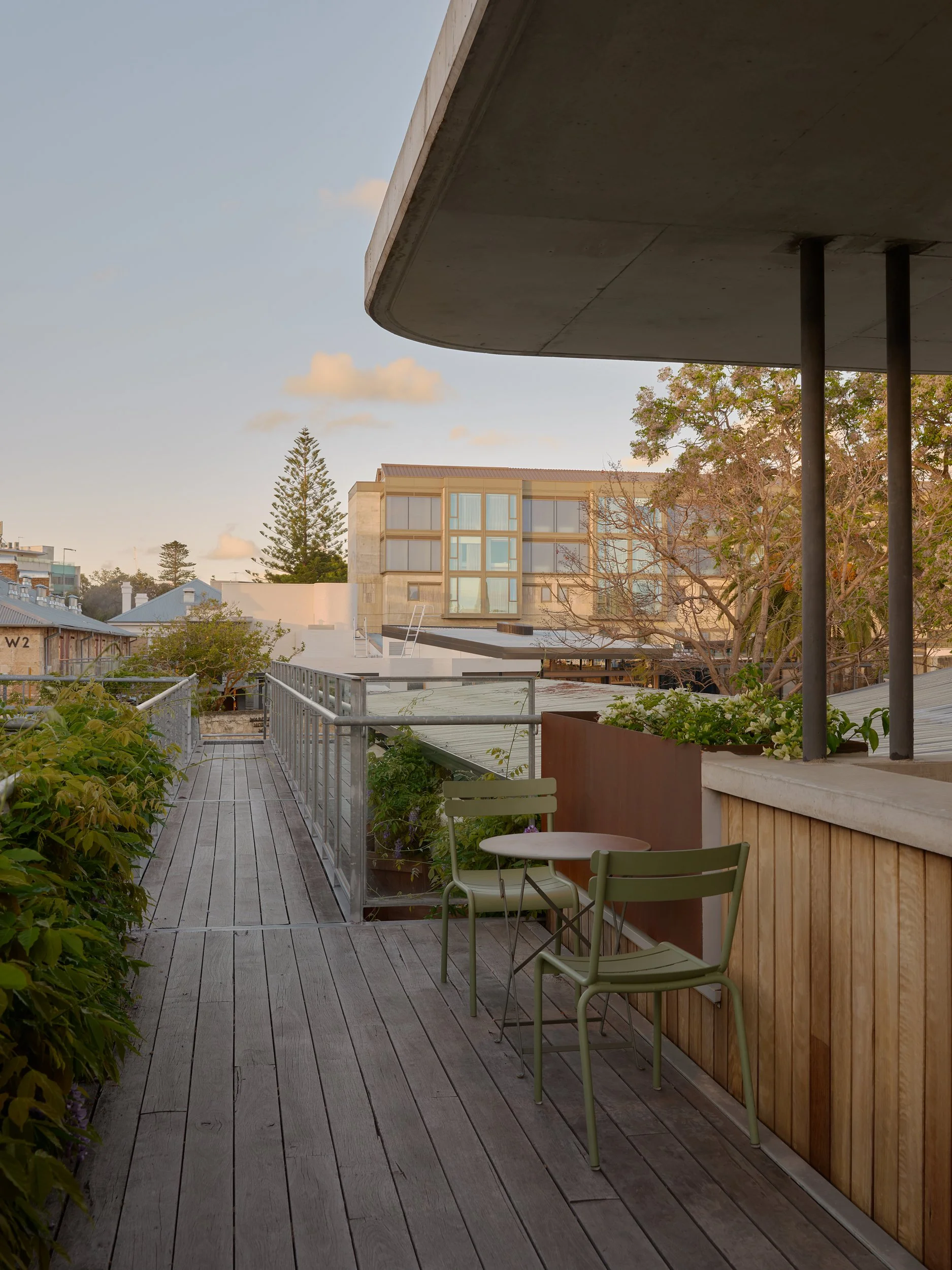 View of a balcony with two green chairs and a small table, overlooking neighboring buildings and trees during sunset.