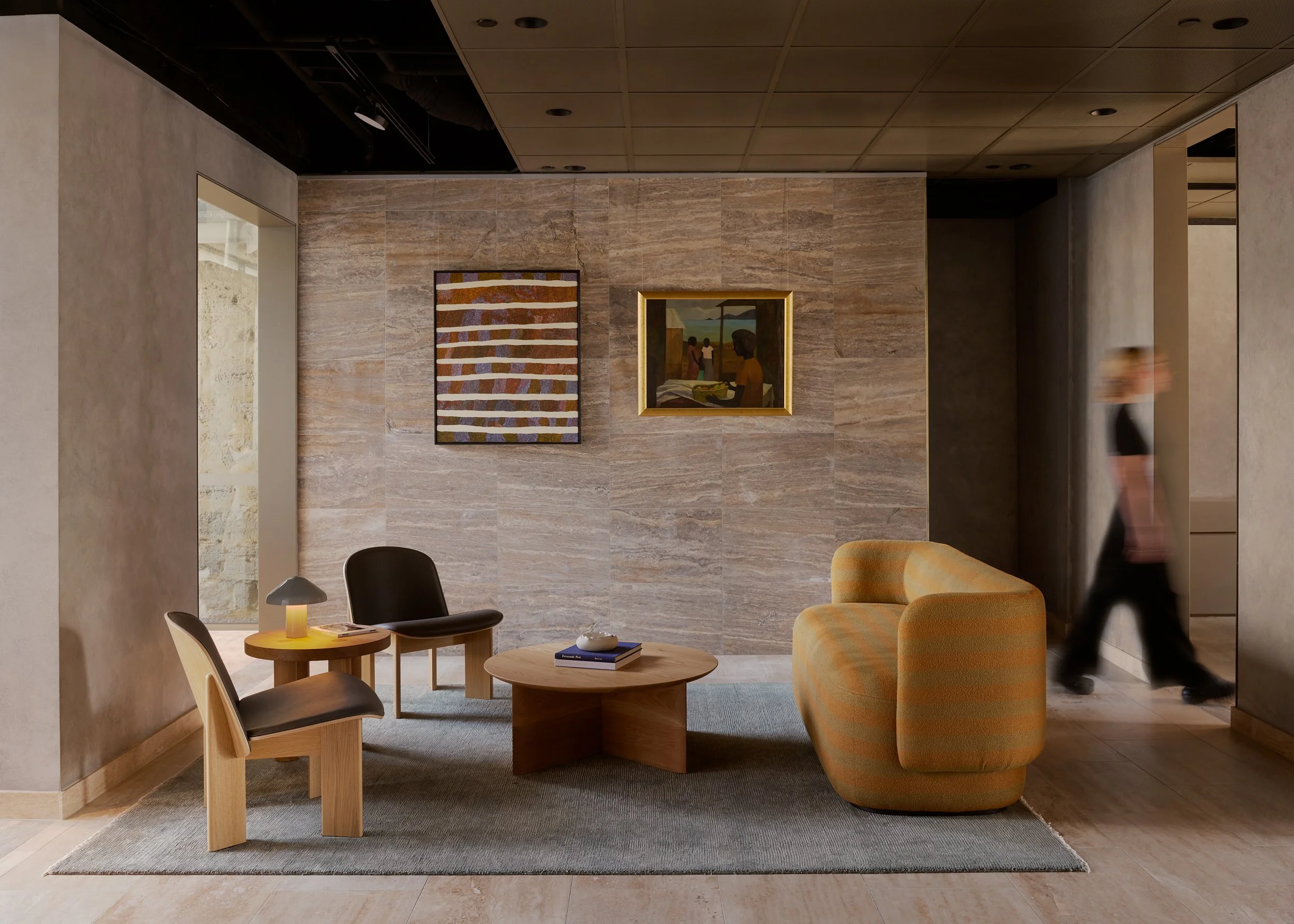 Modern hotel lobby with a seating area including two wooden chairs, a black chair, a yellow striped armchair, and a round wooden coffee table with books and a decorative object. The background features a textured beige wall with two artworks, and a b