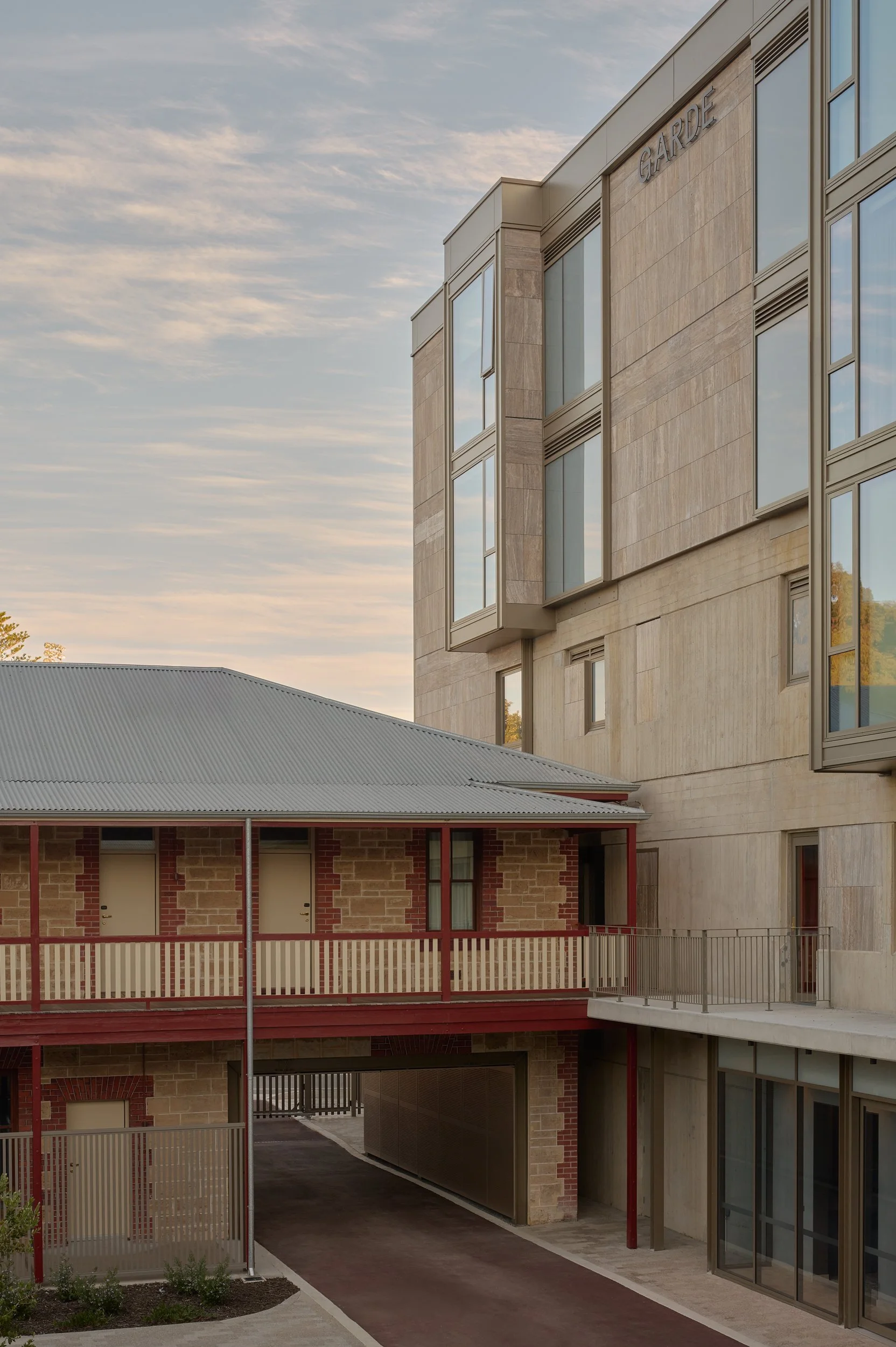 The image shows a modern multi-story building with large windows and stone facade, with a section labeled 'GARDEN'. There is a covered walkway and a parking area underneath, and a bronze railing surrounding the building.