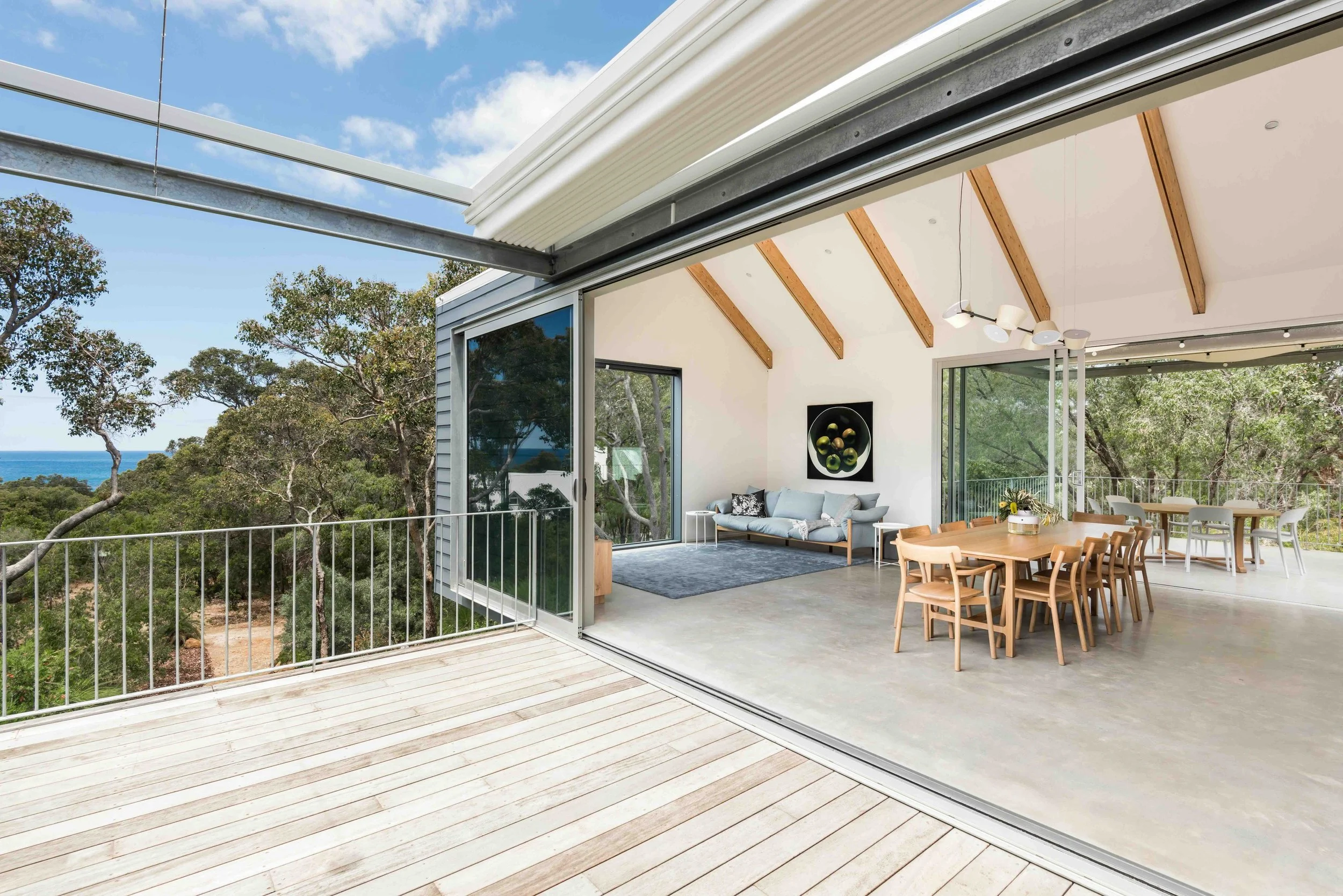Open-concept living and dining area with large glass sliding doors, wooden ceiling beams, contemporary chandelier, and outdoor balcony with trees and ocean view.