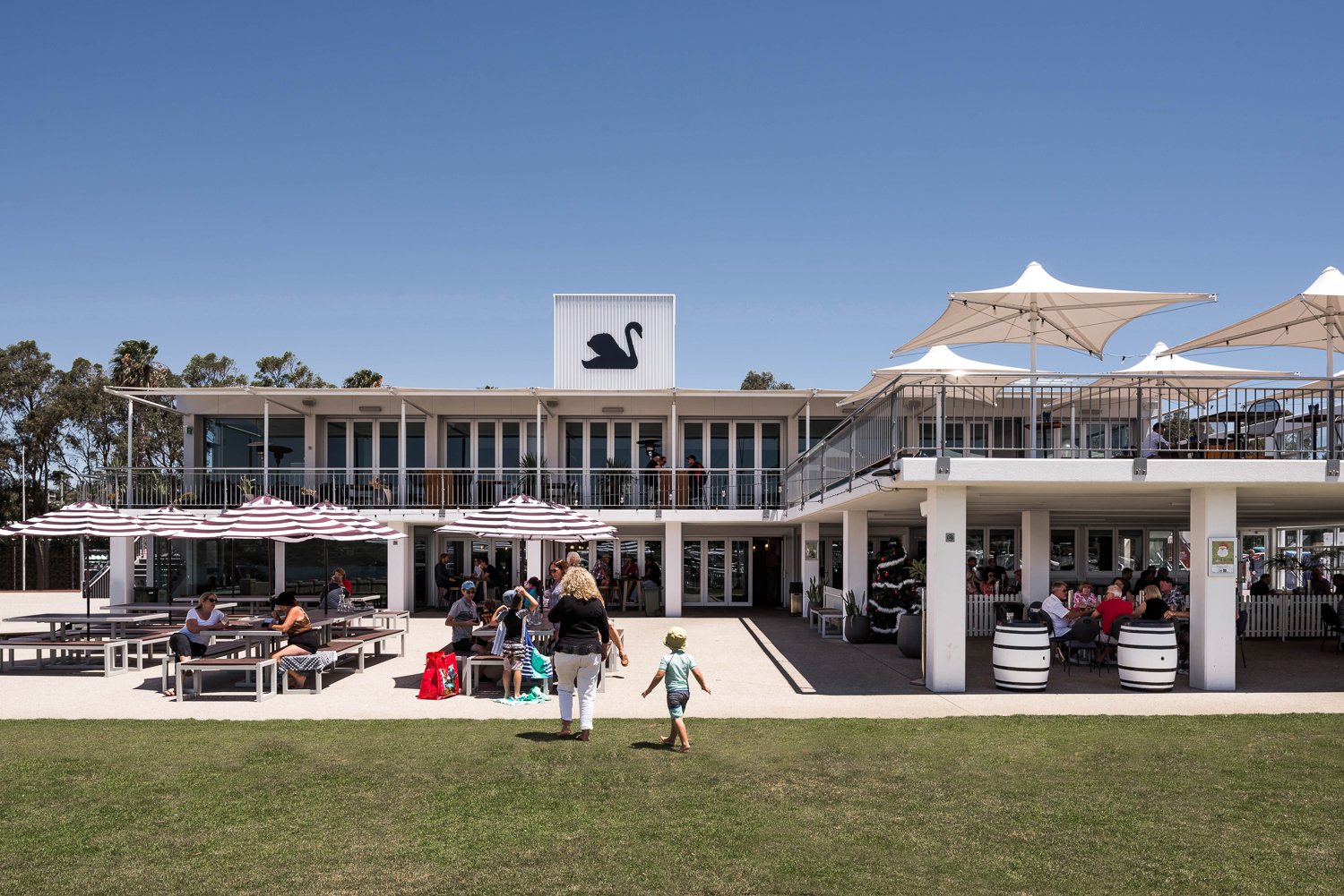 Outdoor restaurant with people sitting at picnic tables and under umbrellas, a second-floor balcony with additional seating, and a black swan logo on the building's roof under a clear blue sky.