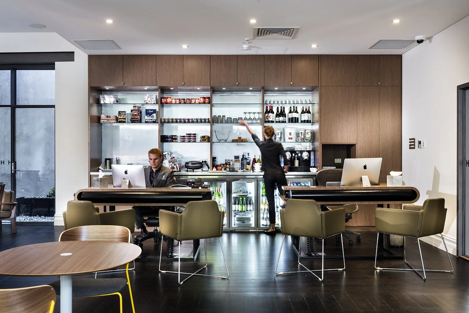 Modern office lounge with a bar area, two people working on computers, and a woman reaching for items on shelves behind the bar.