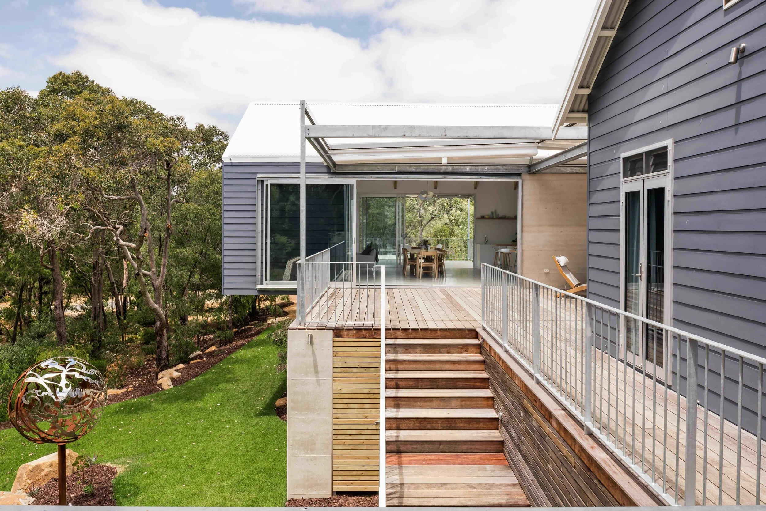 Modern house with outdoor deck, stairs, and railing, surrounded by green lawn and trees.