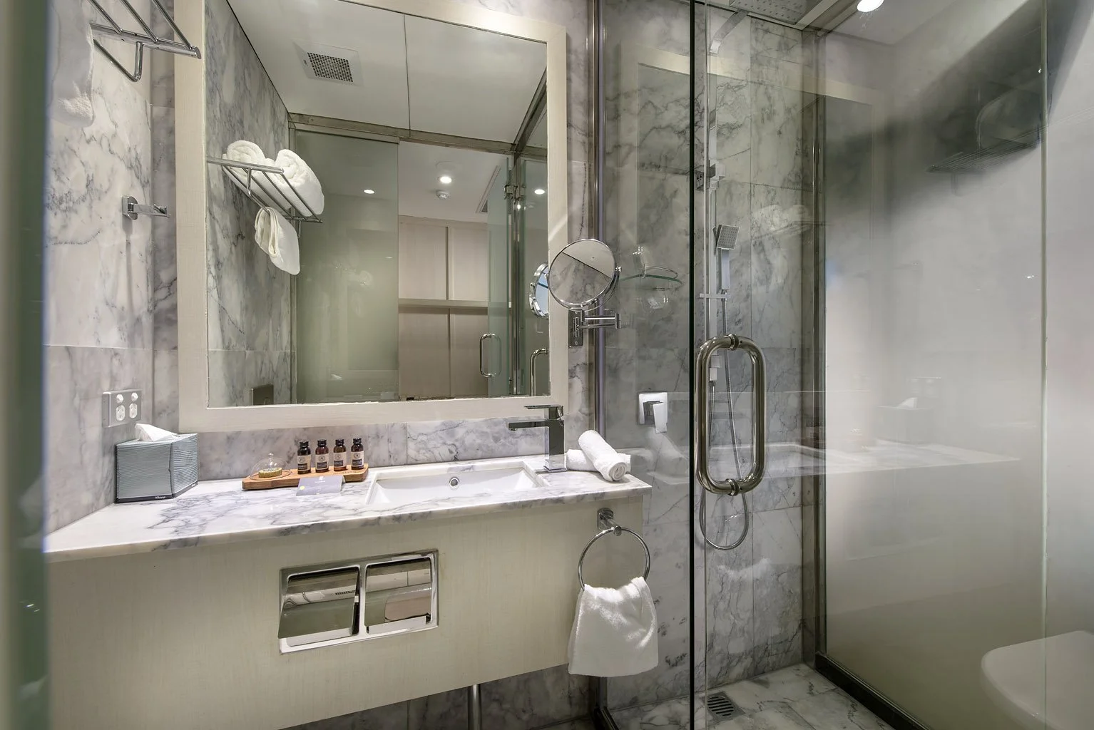 Luxurious bathroom with marble walls and countertop, mirror, toiletries, towels, and a walk-in shower with glass door.