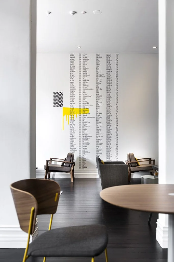 Interior view of a modern lounge or waiting area with chairs and a large wall listing names or information, featuring minimalistic art with yellow paint drip detail.