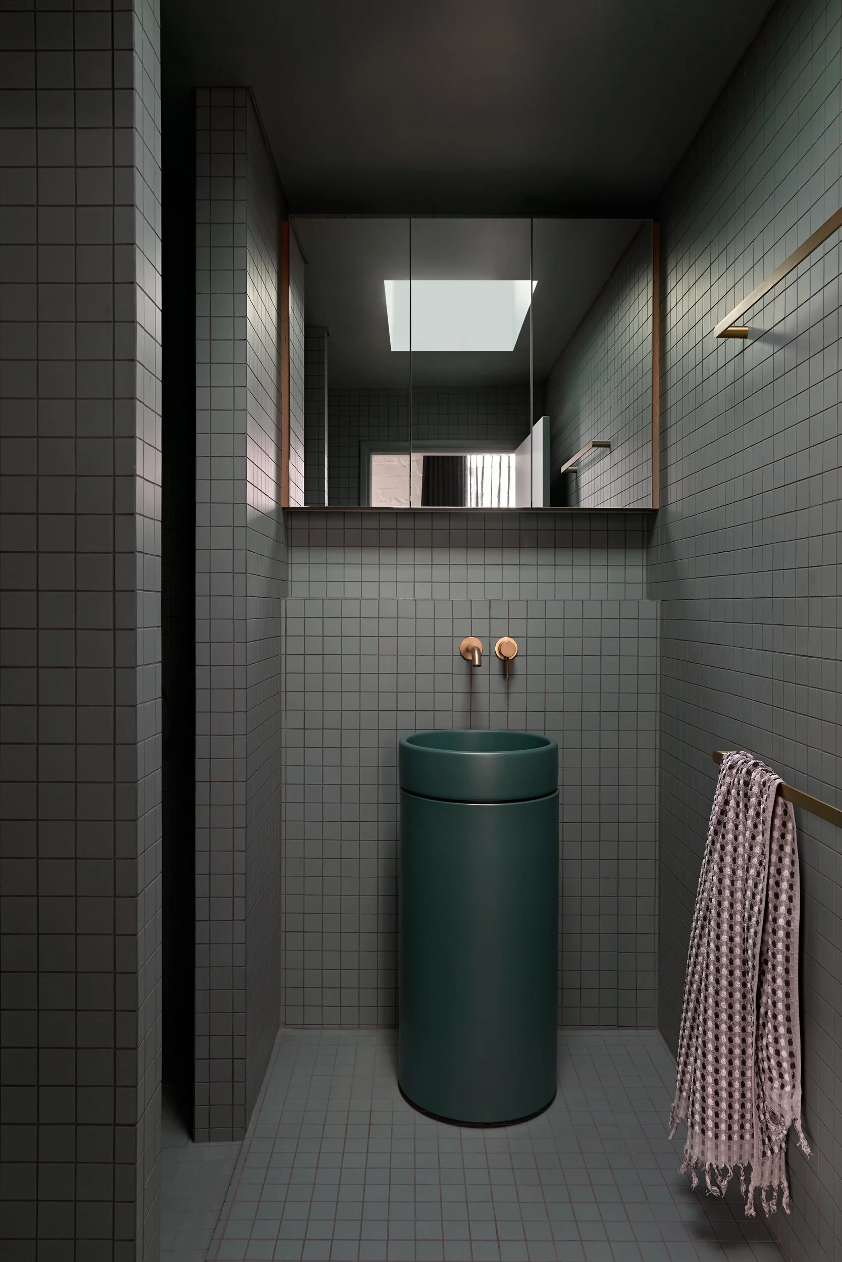 Modern bathroom with a green cylindrical sink, a large mirror, rose gold fixtures, tiled walls and floor, a skylight, and a towel hanging on a gold towel bar.