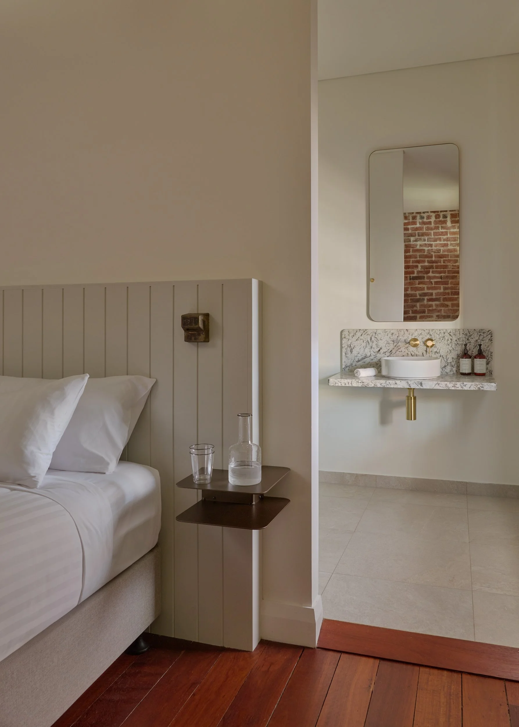 A bedroom with a white bed, bedside table with a glass water pitcher and glass, and a view into a bathroom with a marble sink, mirror, and exposed brick wall.