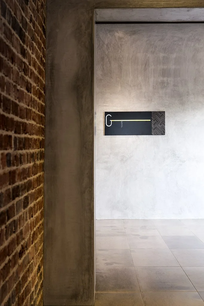 A minimalist interior corner with a brick wall on the left, a concrete wall on the right, and a sign on the back wall displaying a large letter 'G' with a yellow line and the words 'Level G' in small text.