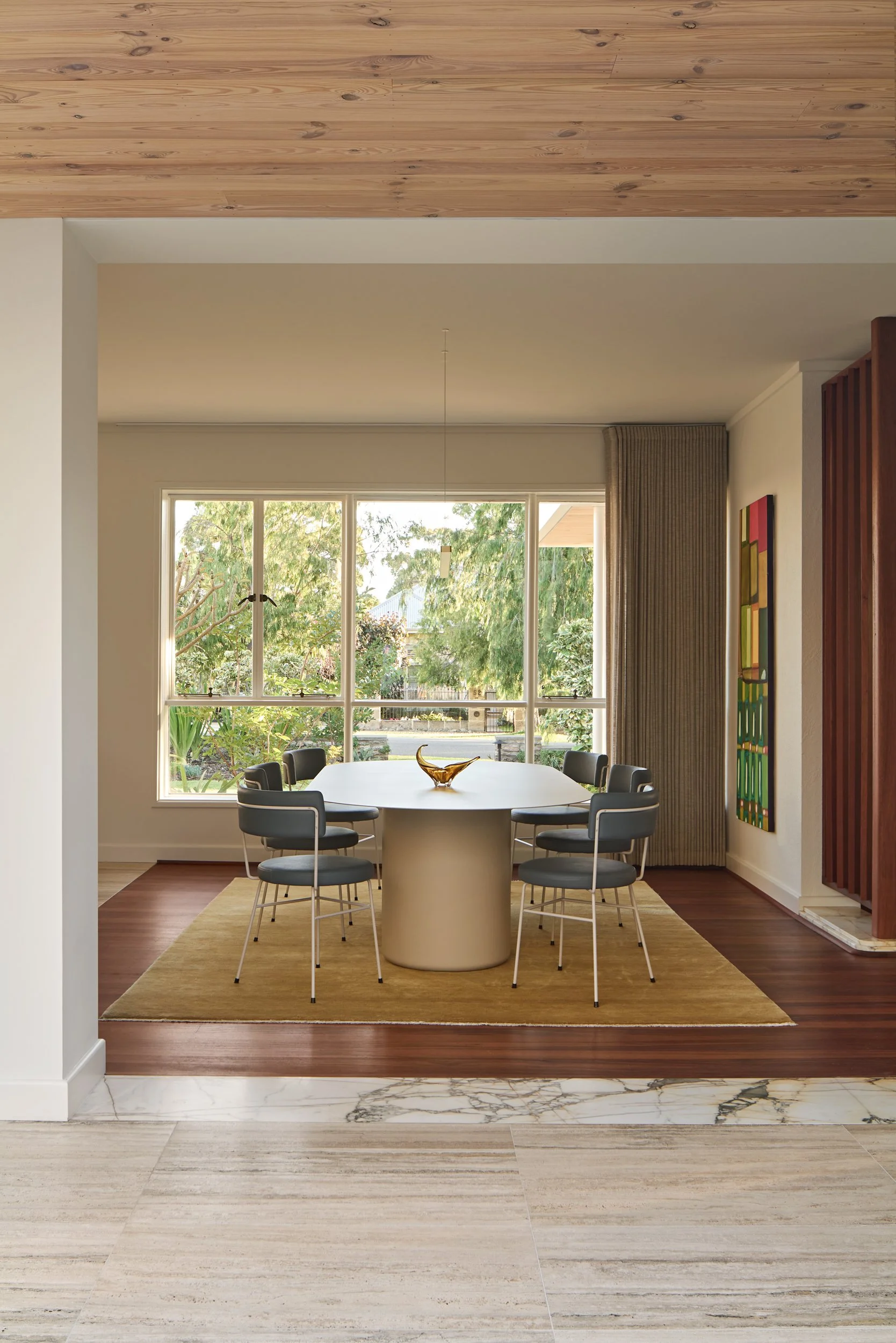 A dining room with a large window overlooking a green yard, featuring a white oval table surrounded by black chairs, a yellow rug, and artwork on the wall.