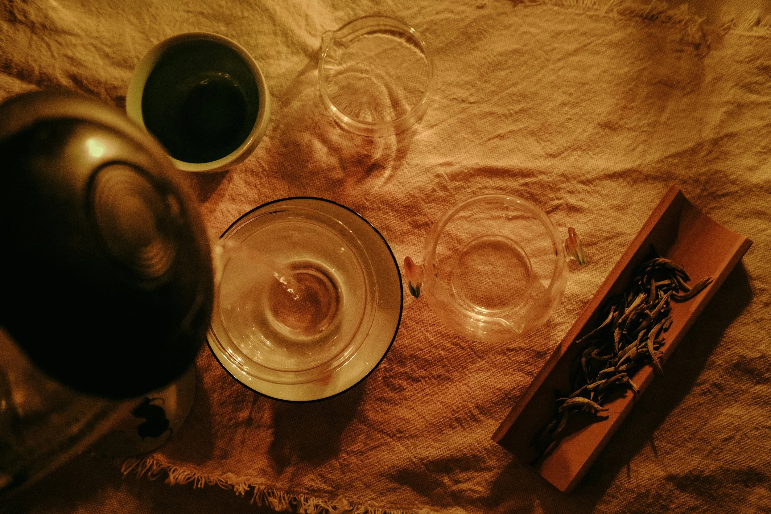 Bottle pouring a liquid into a glass with two other glasses and a wooden tray of dried tea leaves on a beige cloth surface under warm lighting.