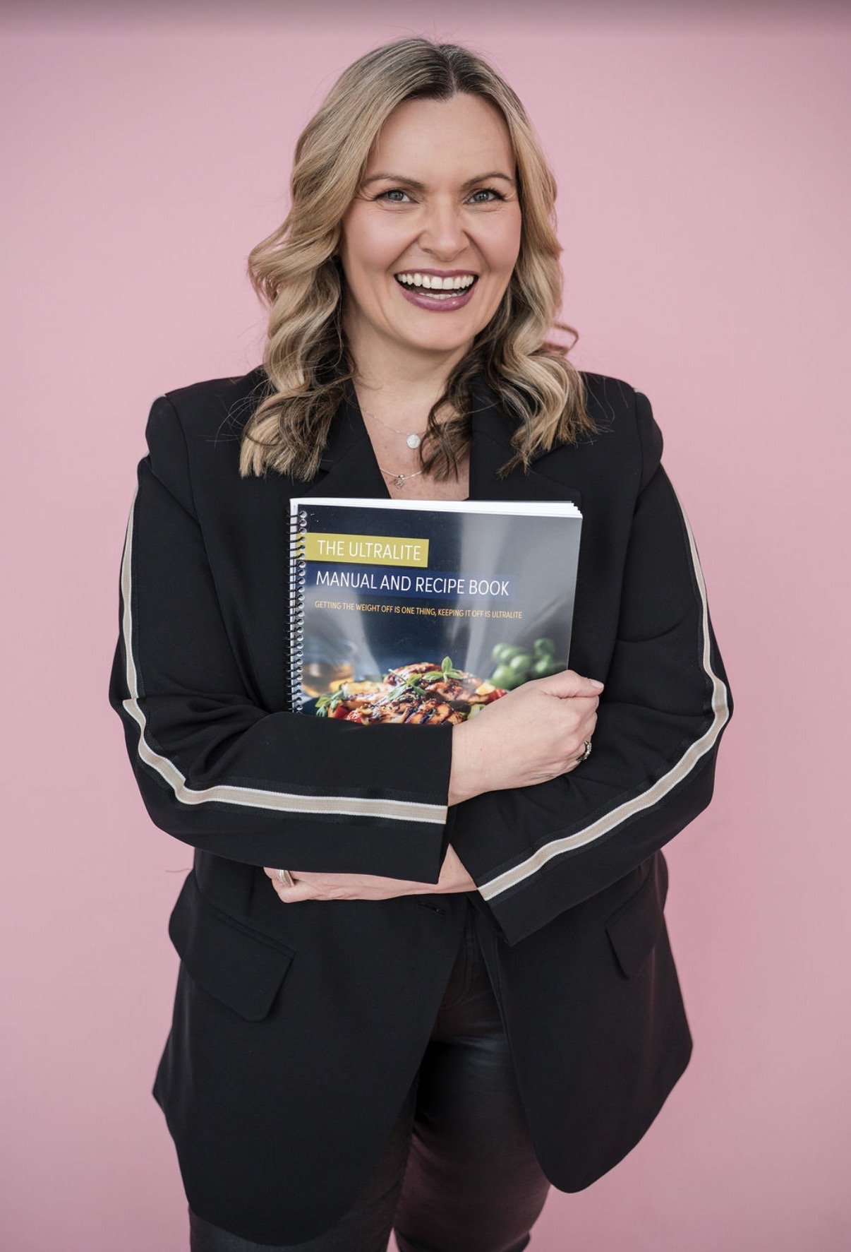 A woman smiling and holding a cookbook titled 'The Ultralite Manual and Recipe Book' against a pink background.