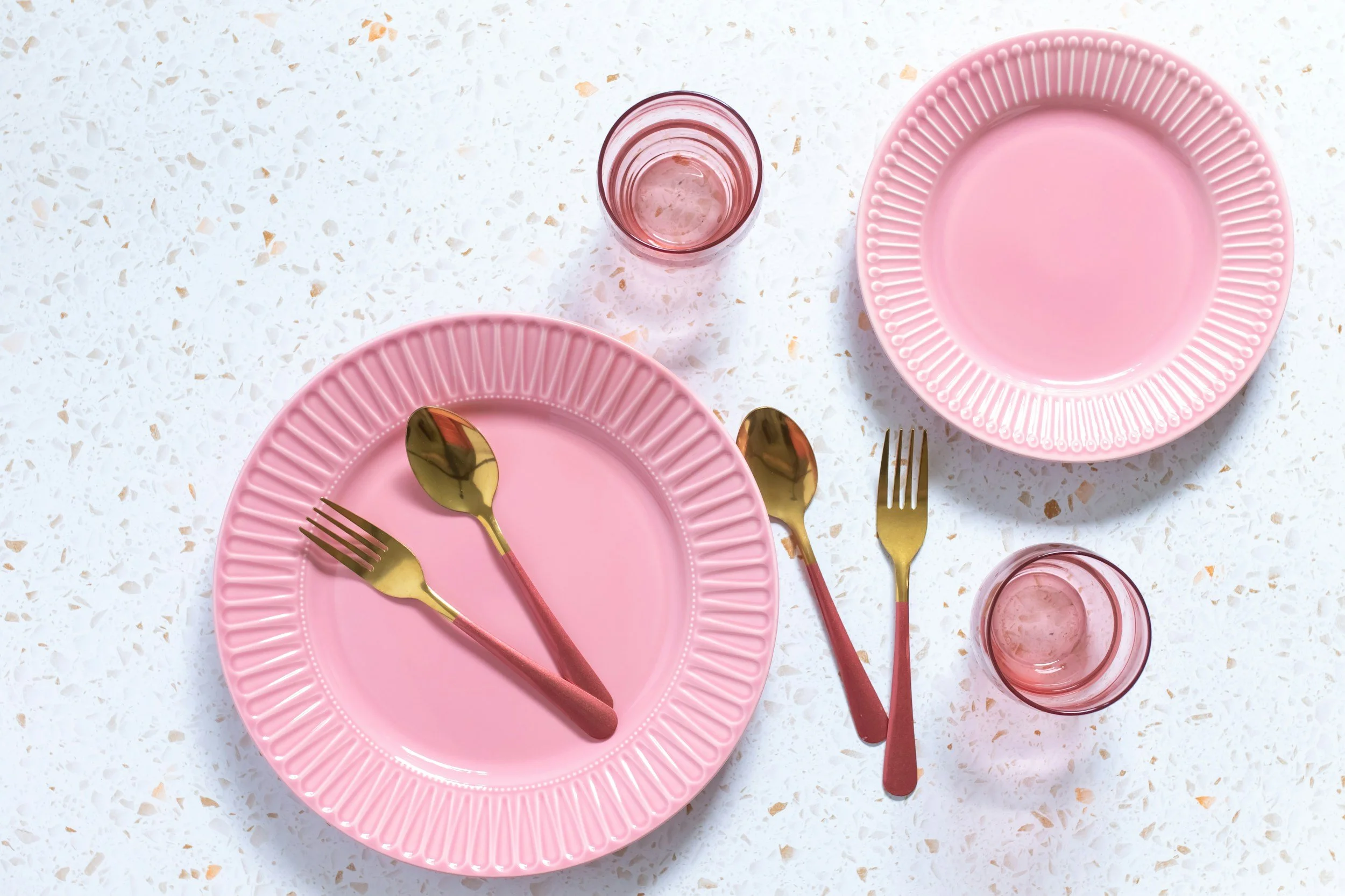 Pink plates and gold utensils set on white countertop with two pink glasses.