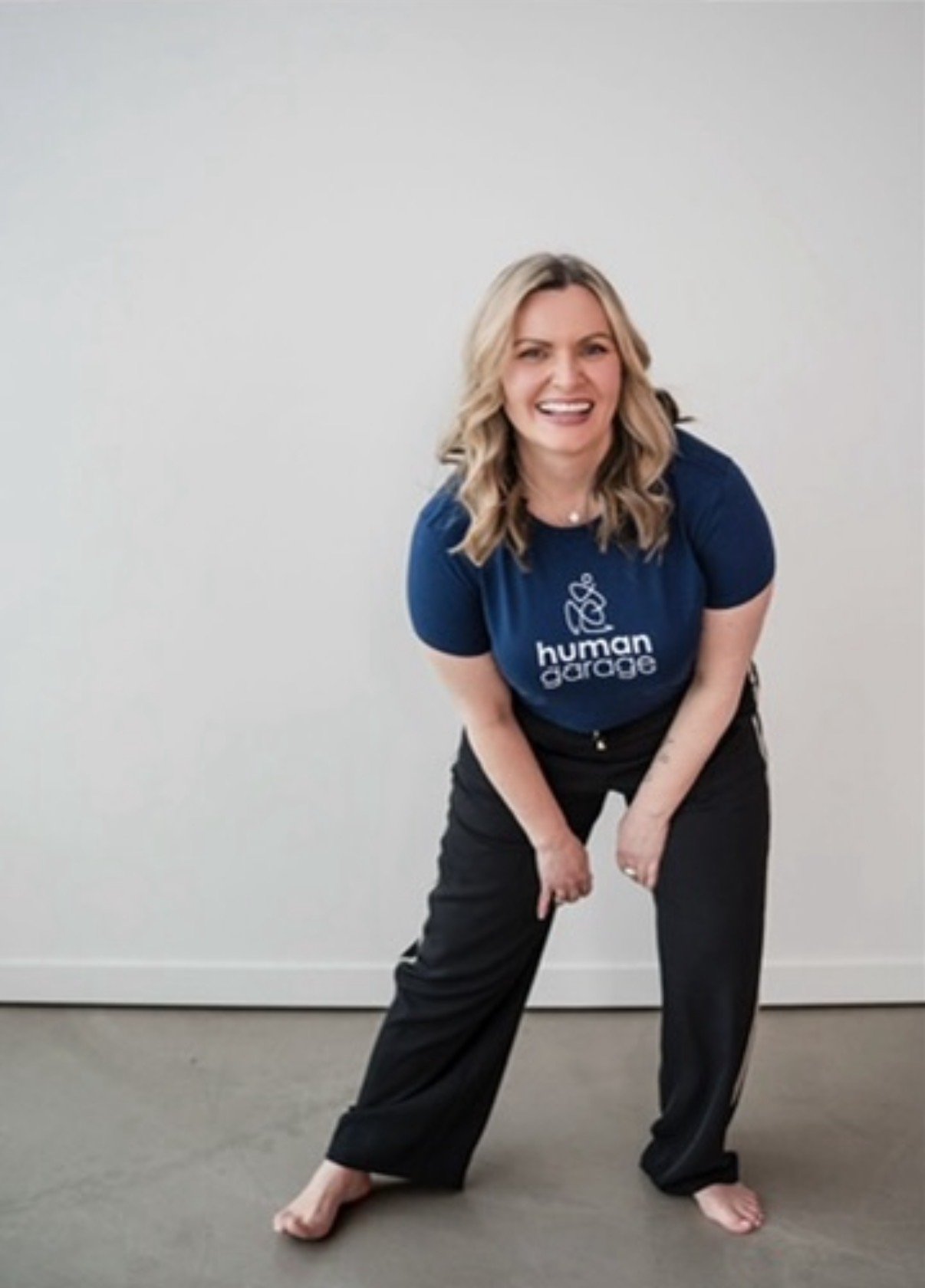 A woman with blonde hair wearing a dark blue t-shirt with the words 'human darge' and a logo, black pants, and no shoes, smiling and leaning forward with her hands on her knees against a plain white wall.