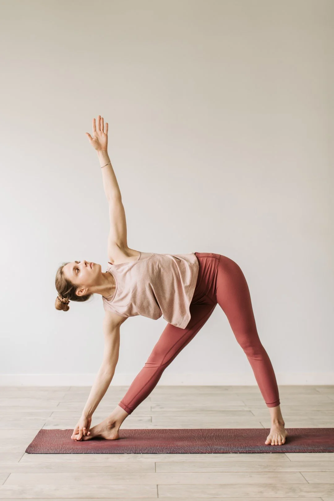 Woman performing yoga pose on a red mat in a minimalistic room with light hardwood floors and plain light gray wall.