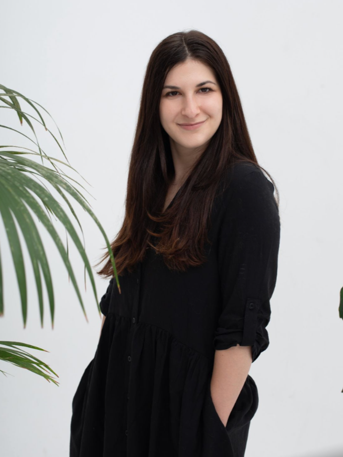 A young woman with long brown hair, wearing a black dress or shirt, standing in front of a plain white wall with green plants on the sides, smiling softly at the camera.