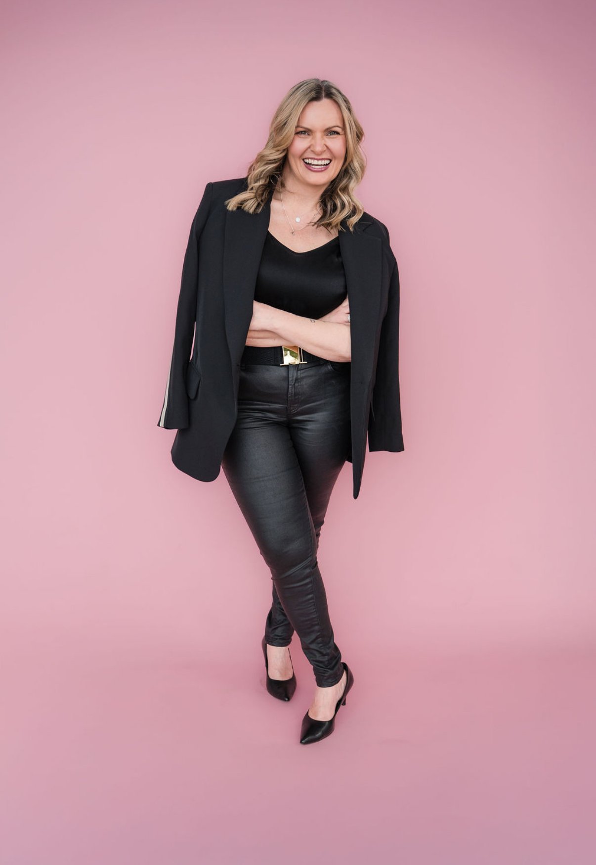 A woman with blonde wavy hair, wearing a black blazer over a black top, leather pants, and black high heels, smiling and standing cross-armed against a pink background.
