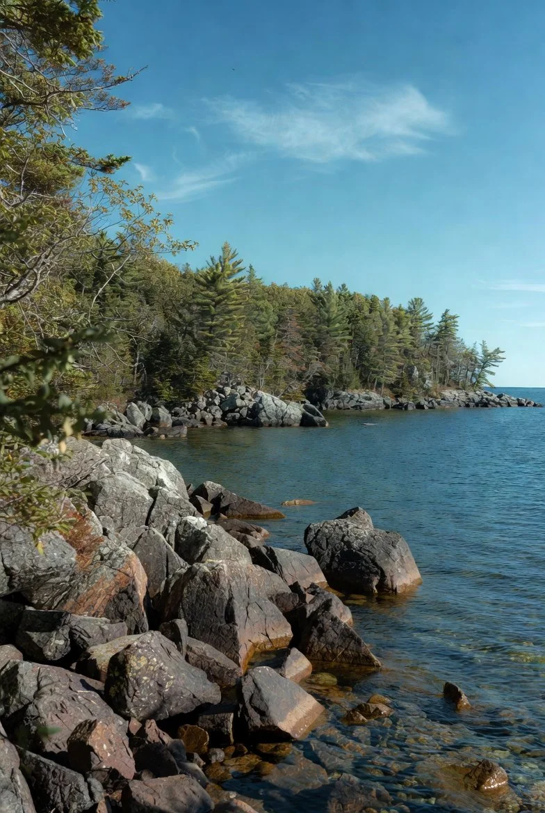 A sailboat with a red hull and white sails sailing on water, with a background of mountains and clouds under a partly cloudy sky.