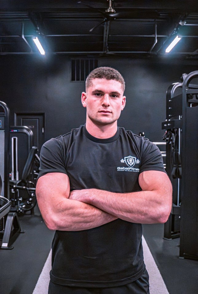 A young man with short brown hair standing with arms crossed in a gym, wearing a black t-shirt with the Gabai Fitness logo, surrounded by gym equipment and dim lighting.