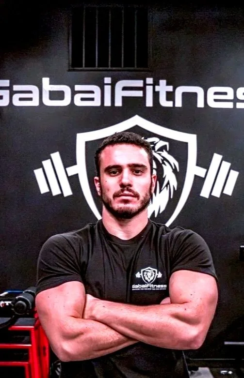 A man with short dark hair and a beard in a black Gabai Fitness shirt crossing his arms in front of a gym wall with a logo of a lion's head within a shield and the word Gabai Fitness.