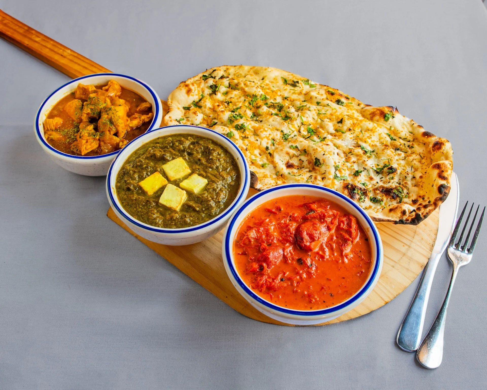 Indian meal with naan bread, three bowls of curry, spoon, and fork on a gray table.