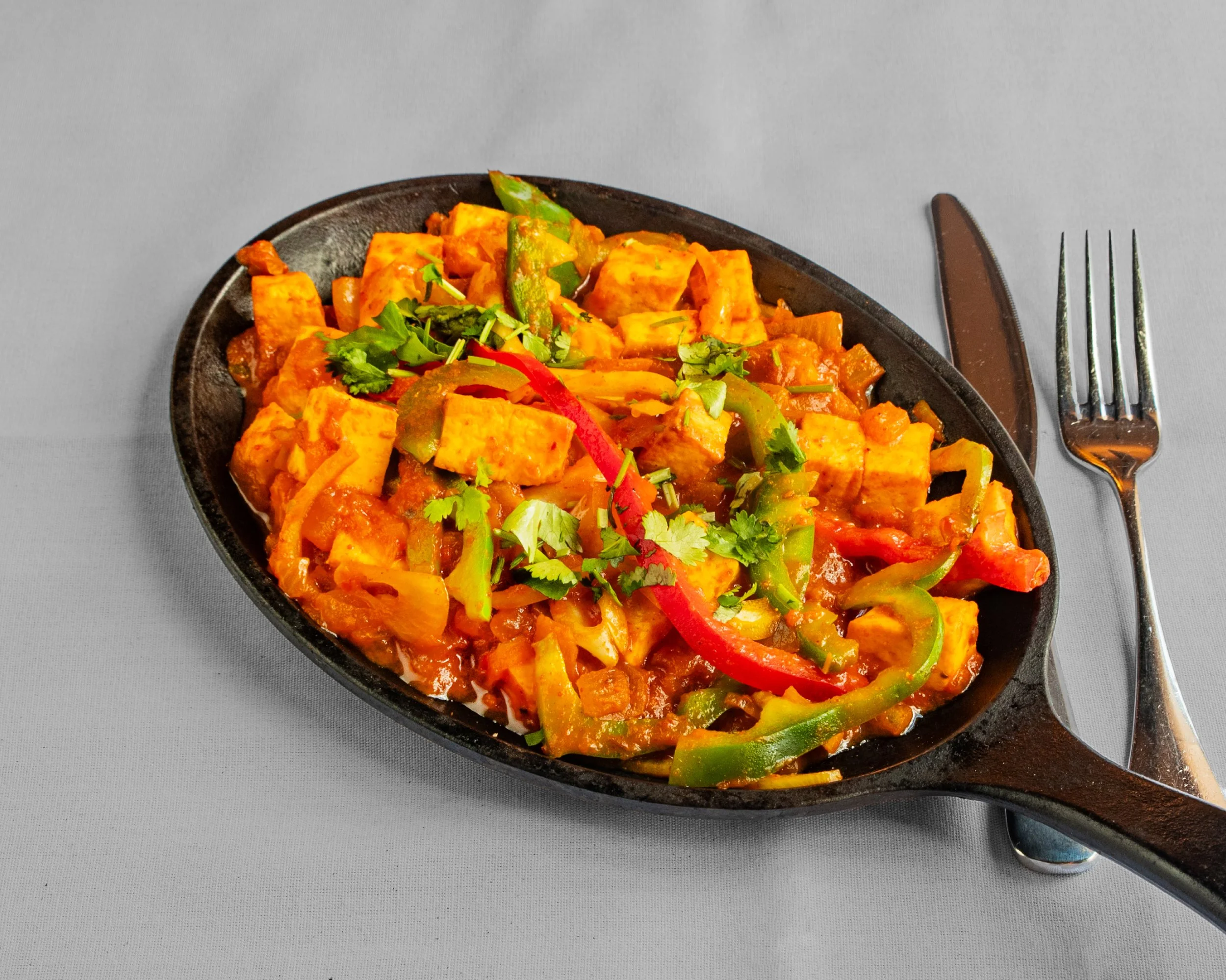 A sizzling dish of tofu and vegetables served in a black cast iron skillet, garnished with fresh cilantro, on a gray tablecloth with a knife and fork beside it.