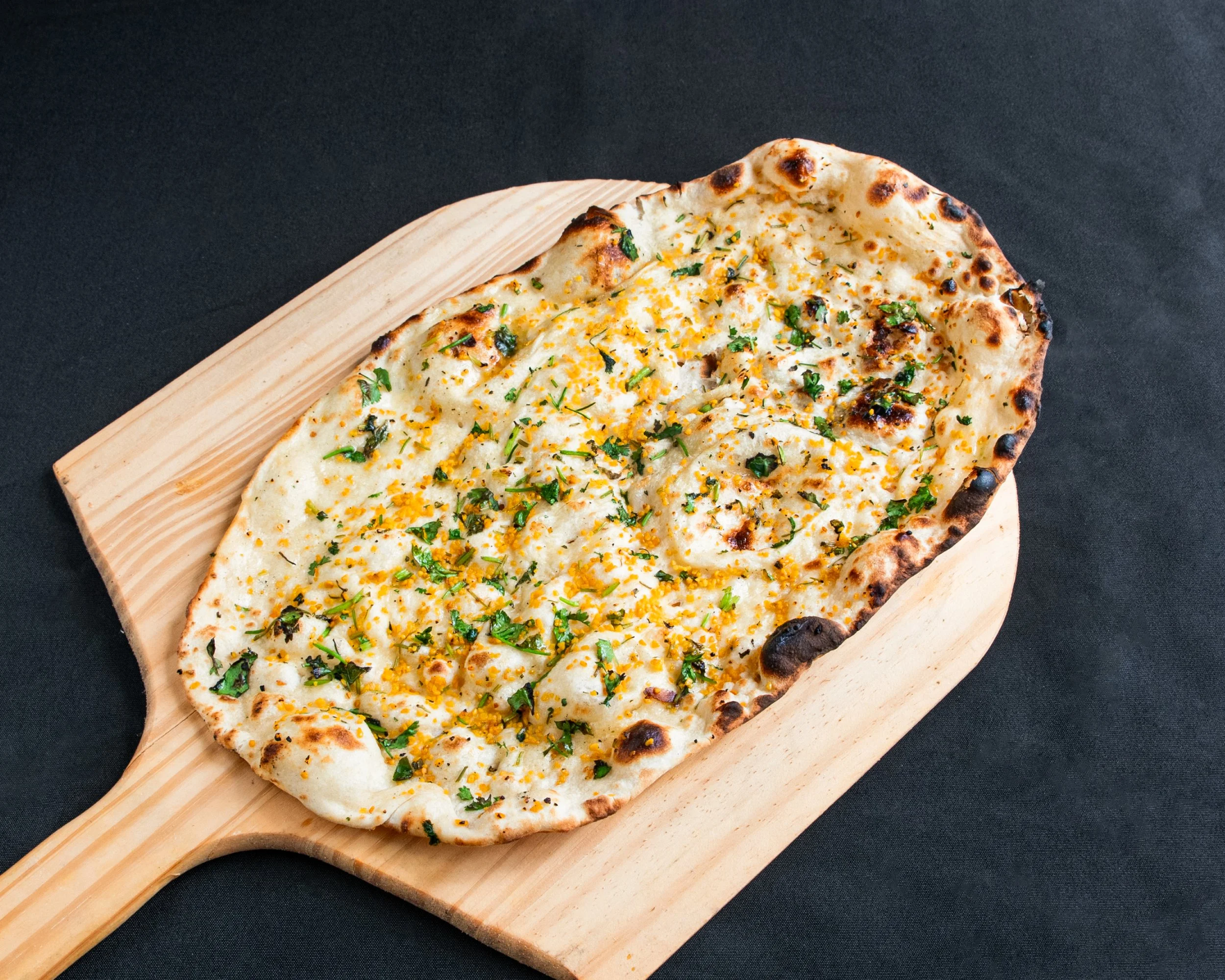 A pizza with a creamy white sauce, sprinkled with herbs and garnished with cheese, on a wooden pizza peel against a dark background.