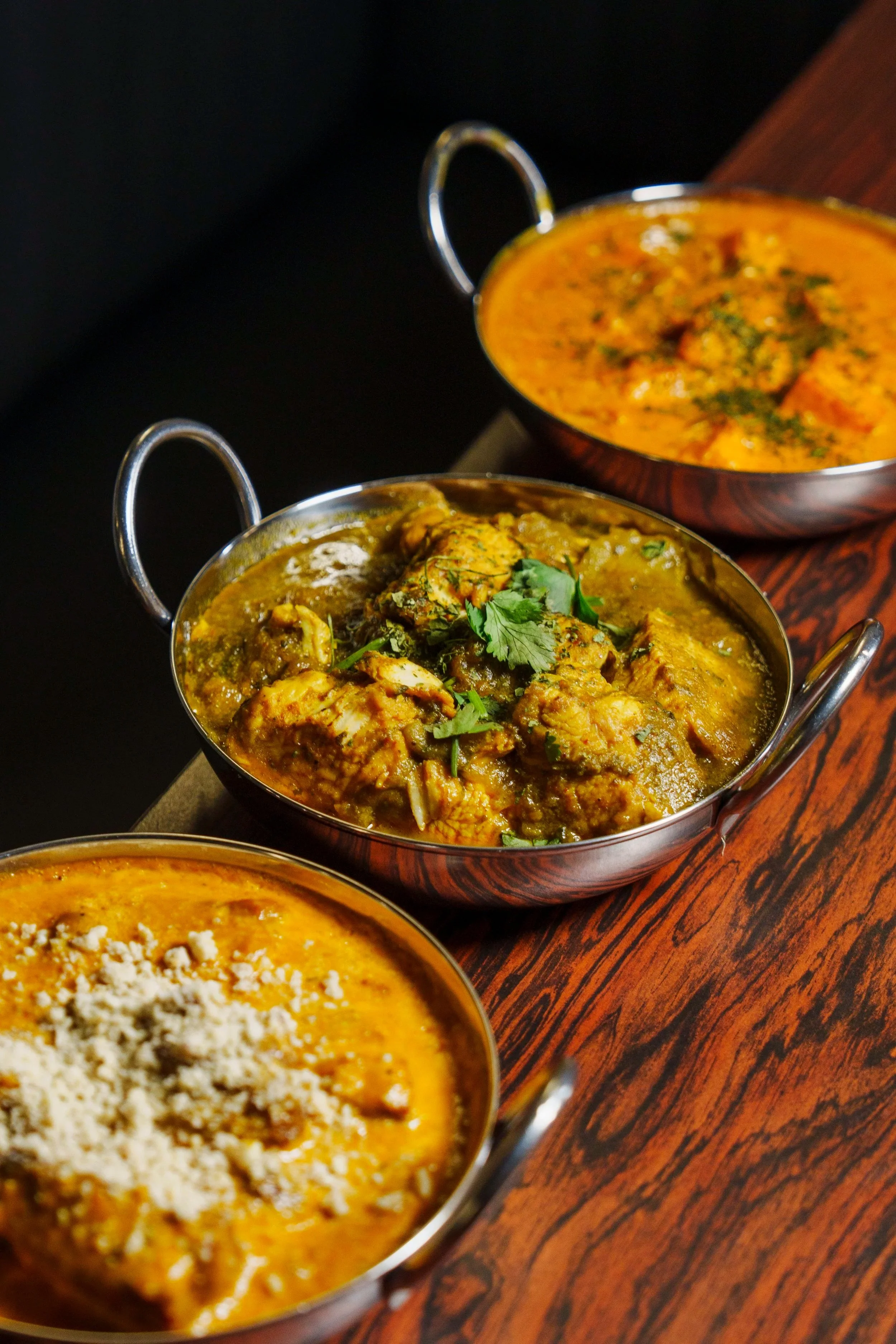 Three bowls of Indian curry dishes on a dark wooden table, with the middle bowl containing chicken curry garnished with cilantro.