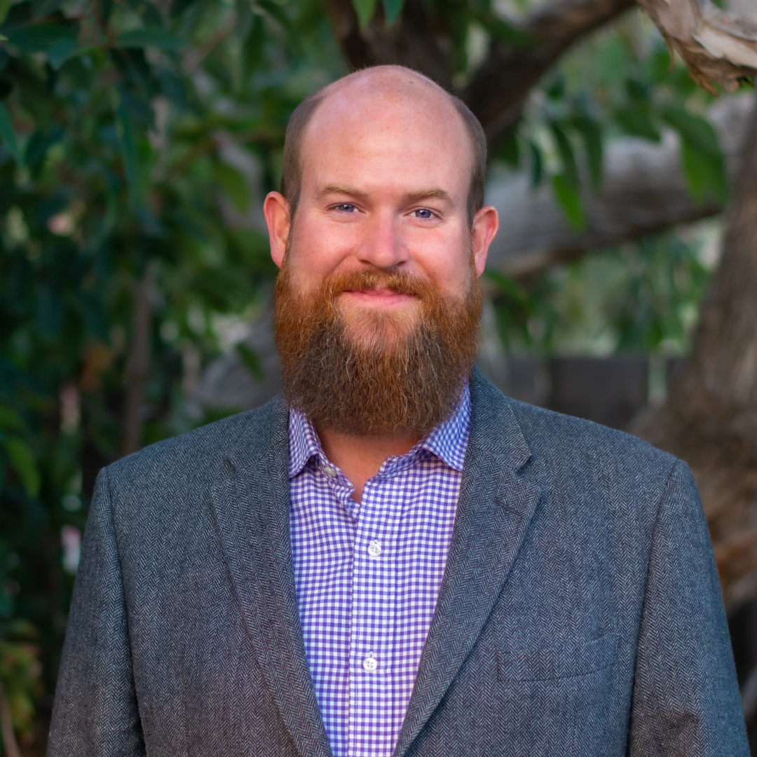 A man with a beard and bald head standing outdoors in front of green foliage, wearing a gray blazer and a purple checkered shirt.