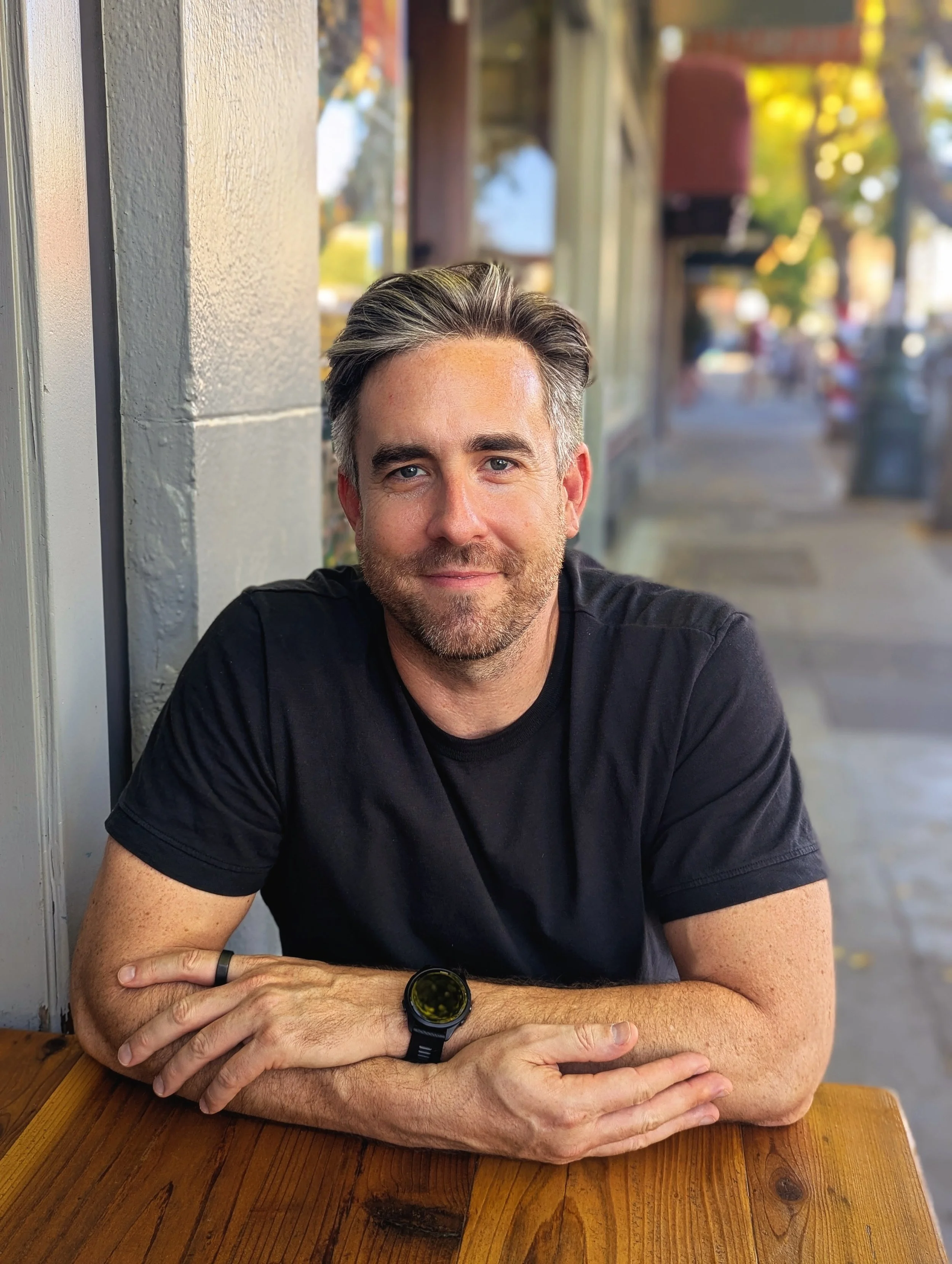 A man with gray hair and a beard sitting at a wooden table outdoors, wearing a black t-shirt and a black watch, smiling at the camera.