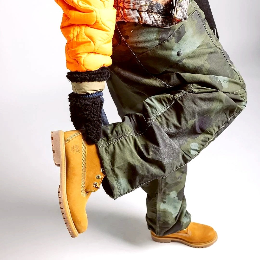 A person wearing tan timberland boots, camouflage cargo pants, an orange puffer jacket, and black and beige gloves, kneeling on one knee.