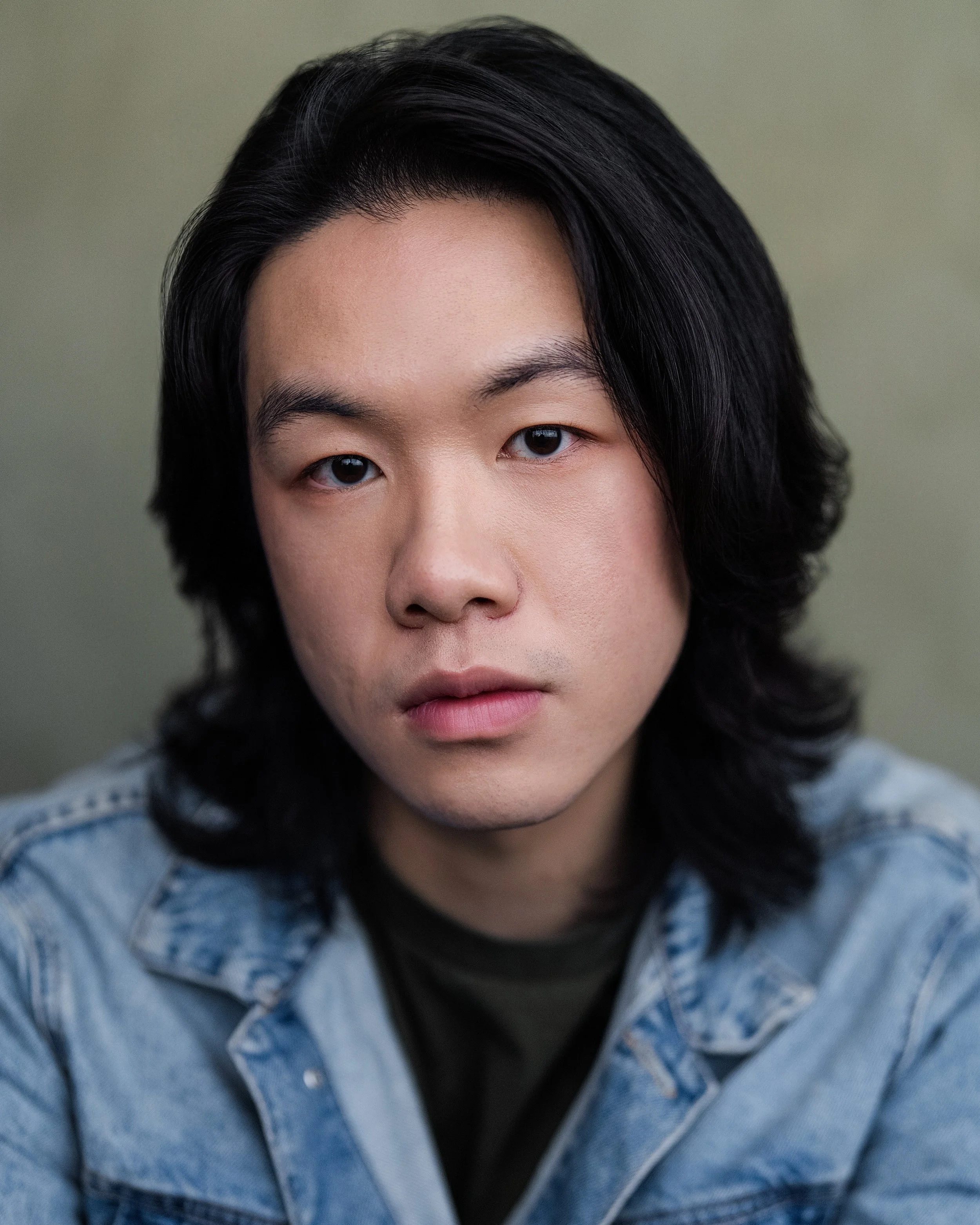 Close-up of a young Asian man with shoulder-length black hair wearing a denim jacket, looking directly at the camera.