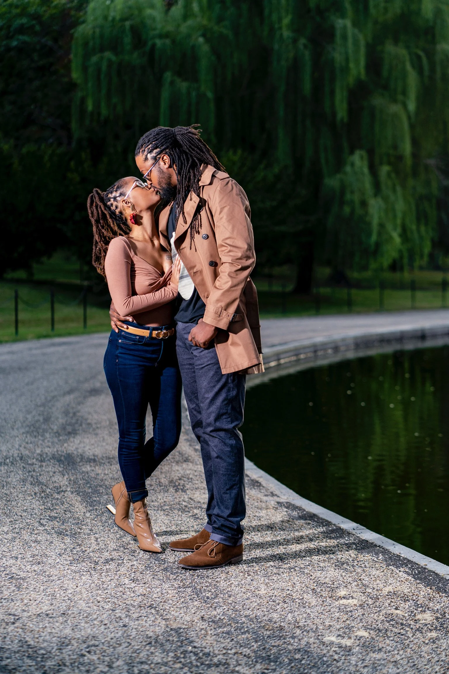 A couple passionately kissing outdoors by a pond in a park, surrounded by green trees.