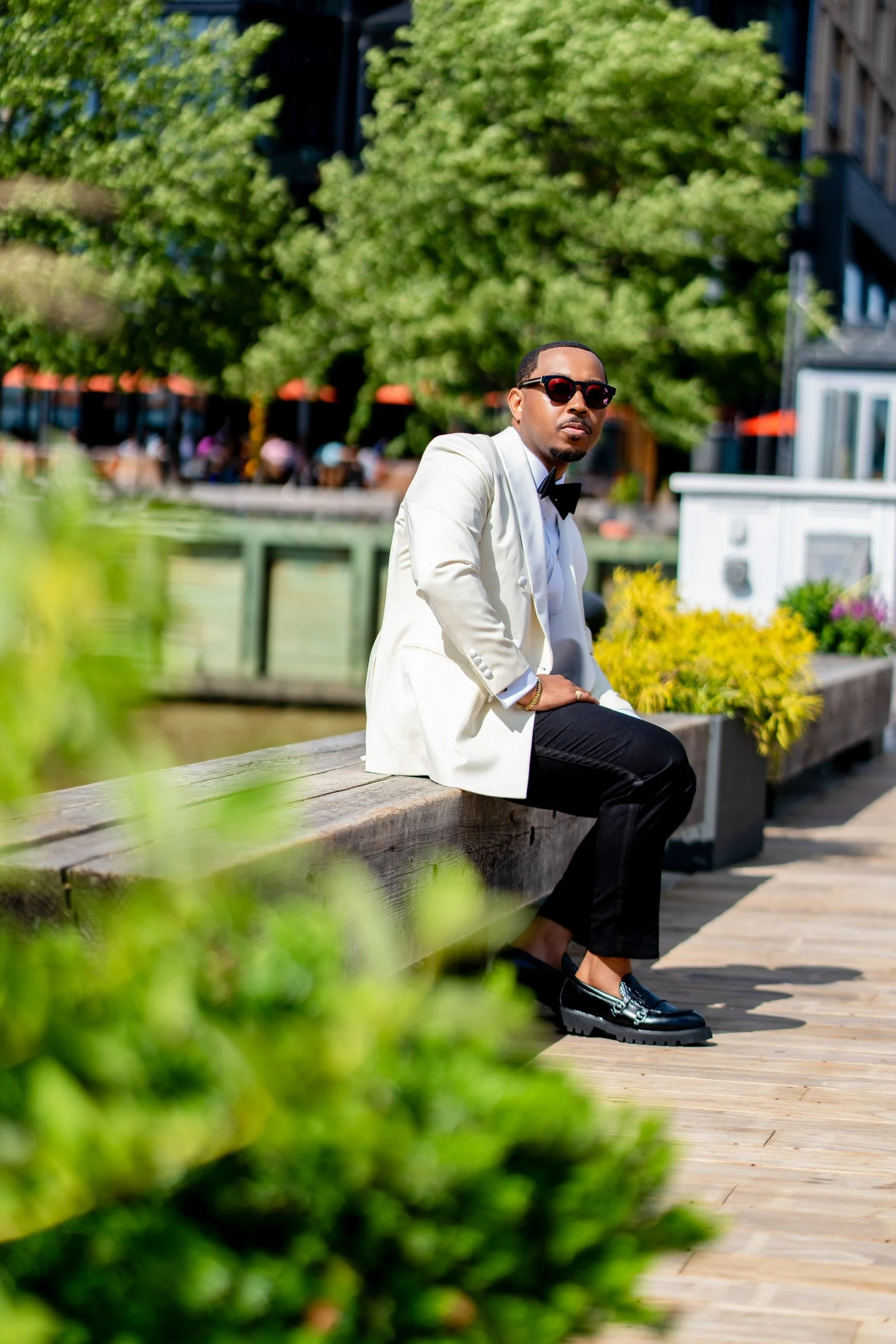 A man in a white tuxedo jacket, black pants, and black shoes sitting on a wooden bench outdoors, wearing sunglasses and a bow tie, with a background of green trees and a cityscape.