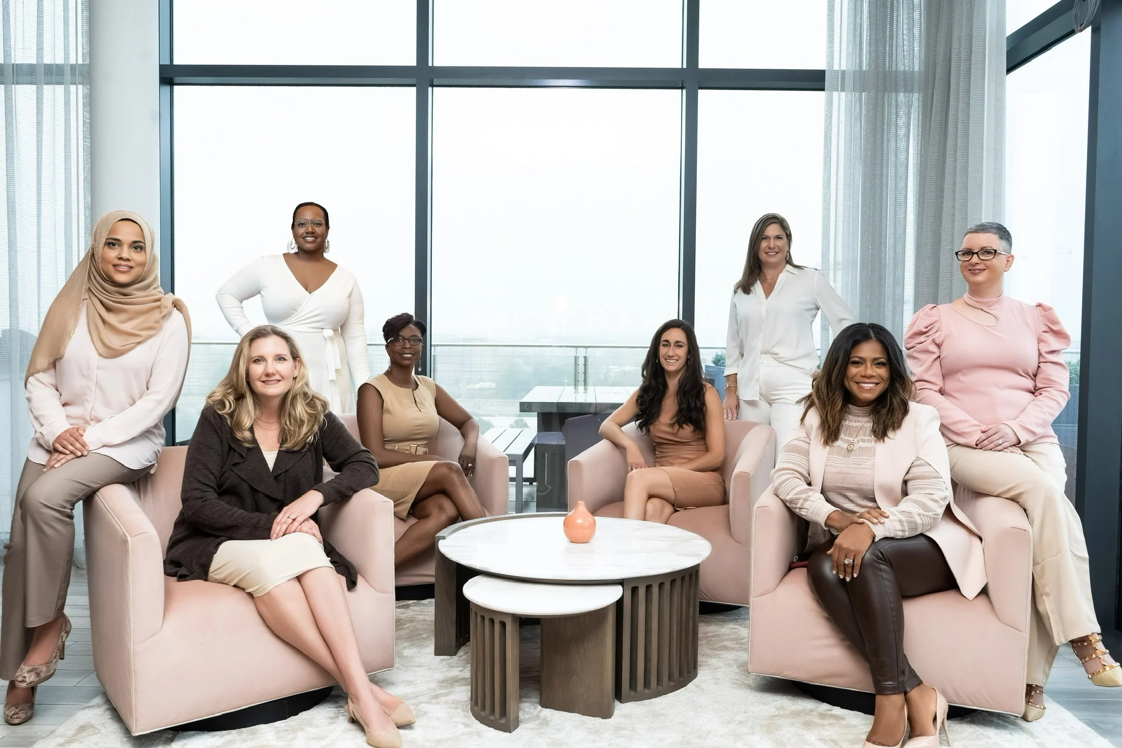 Nine women in stylish business casual attire sitting and standing in a modern, high-rise office lounge with large windows, minimalist furniture, and city view.