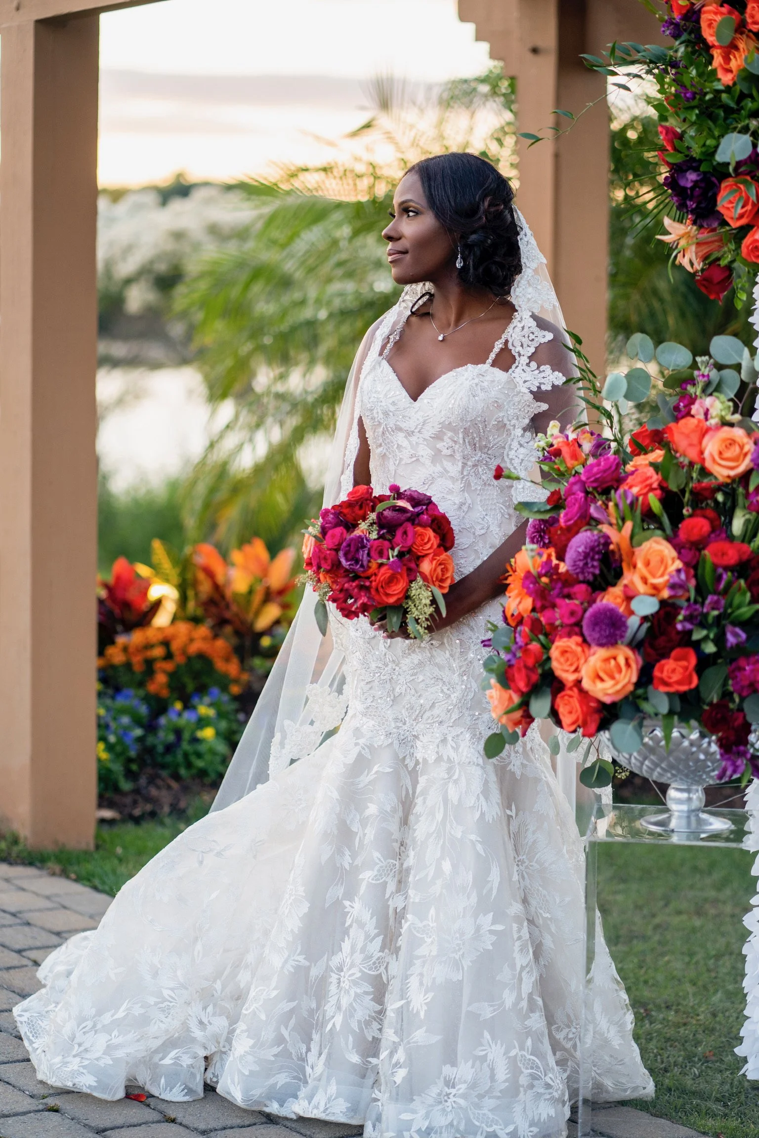 Bride in a white lace wedding dress holding a vibrant bouquet of red, purple, and orange flowers, standing outdoors near a floral arrangement and greenery.
