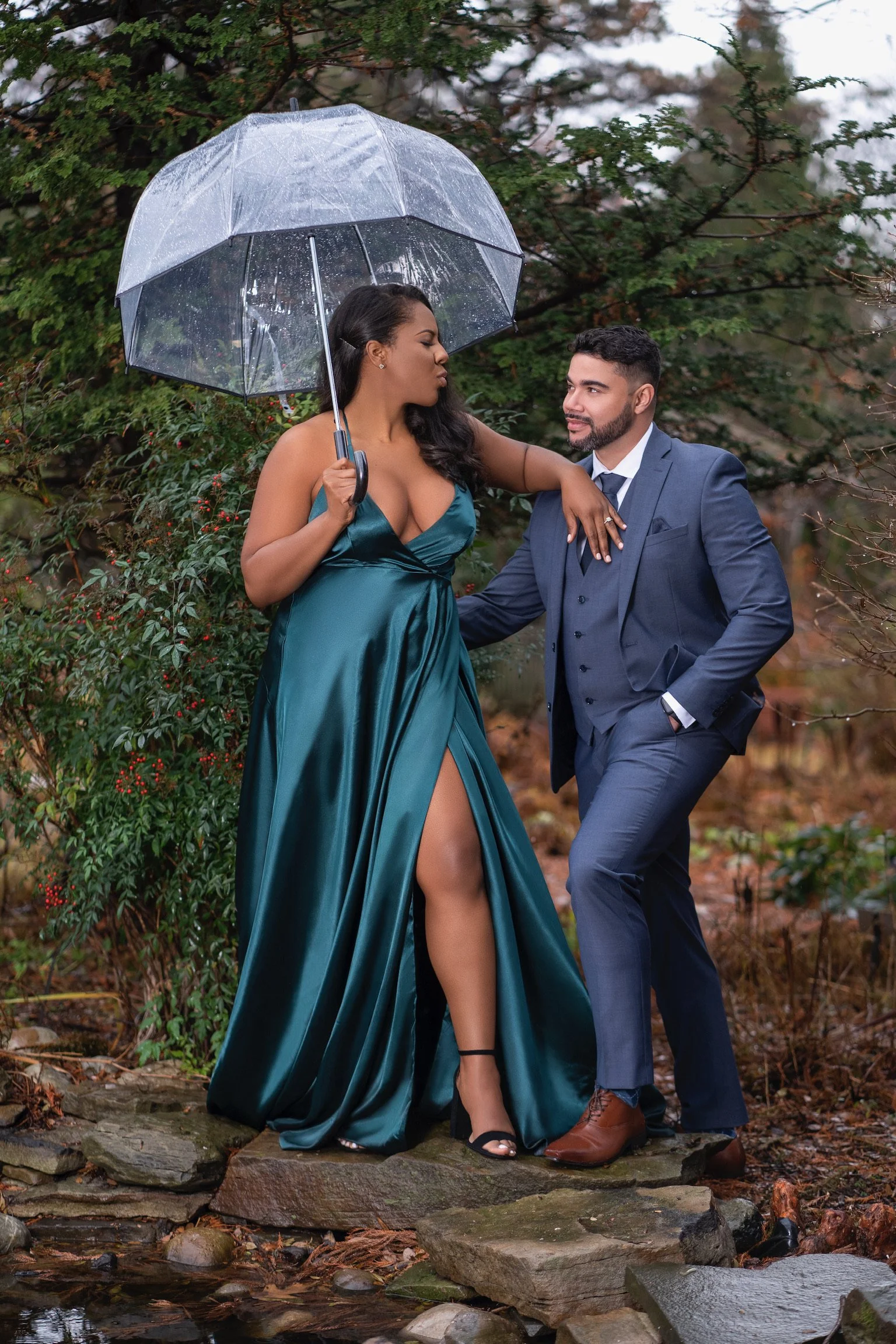 A couple dressed in formal attire standing on rocks outdoors, with the woman holding a transparent umbrella. The woman is wearing a teal gown with a high slit, and the man is in a navy blue suit. They are surrounded by trees and foliage.