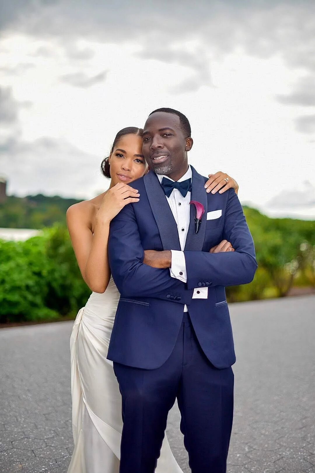 A couple dressed in wedding attire posing outdoors on a cloudy day, with greenery in the background.