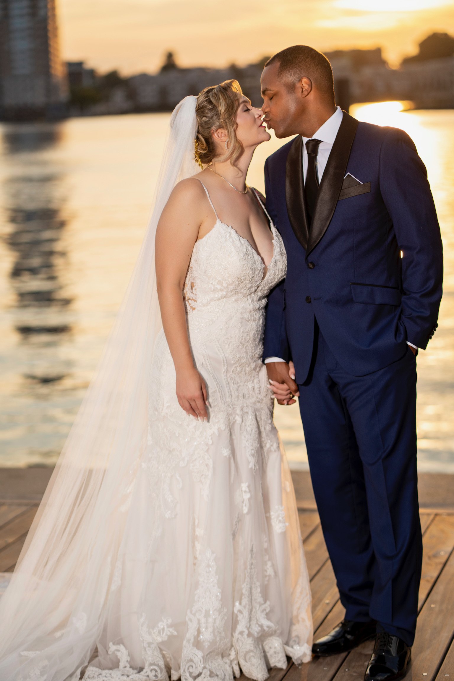A bride and groom share a kiss under the sunset beside a body of water, with city buildings in the background. The bride wears a lace wedding dress and veil, while the groom wears a navy blue tuxedo.