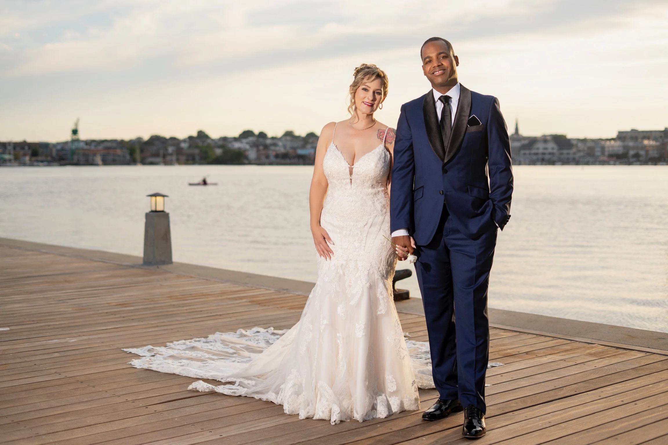 A newlywed couple holding hands on a wooden dock by the water during sunset. The bride is wearing a white lace wedding dress and the groom is in a navy blue tuxedo with black lapels.