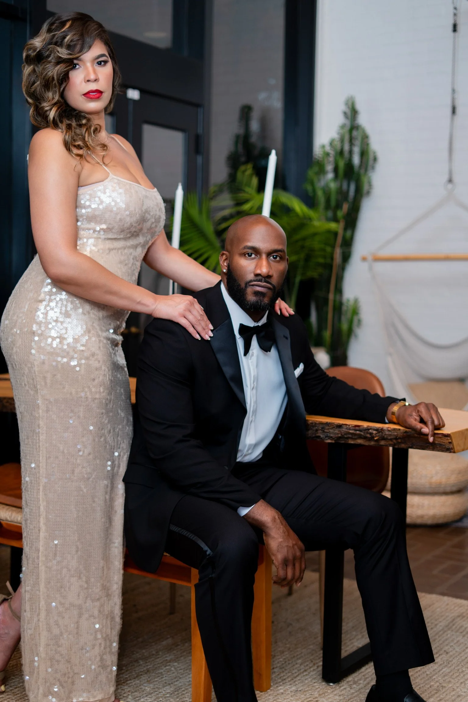 A woman in a sparkling beige gown standing beside a seated man in a tuxedo at an indoor event.