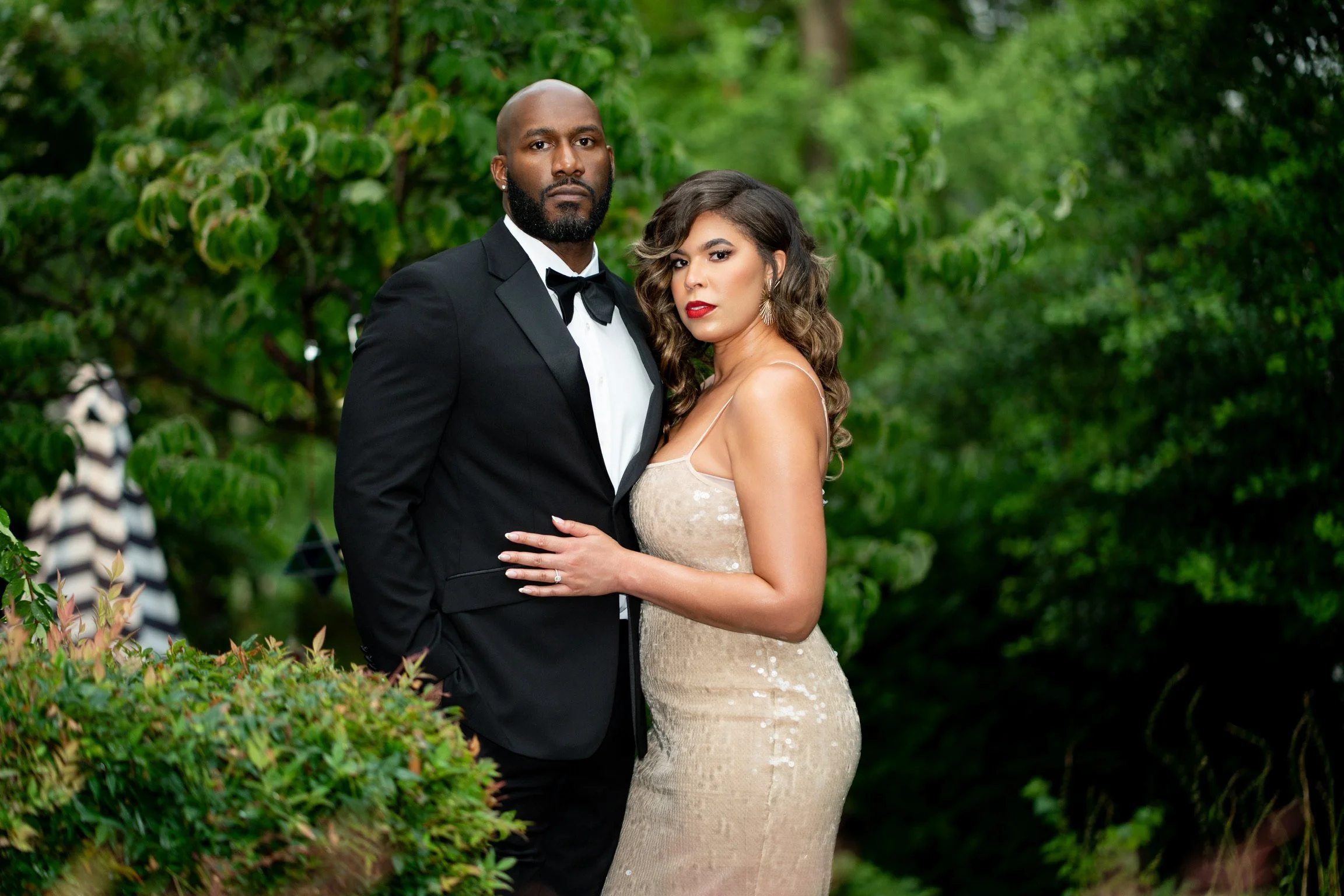 A well-dressed couple poses outdoors surrounded by greenery, the man in a black tuxedo and the woman in a beige evening gown.
