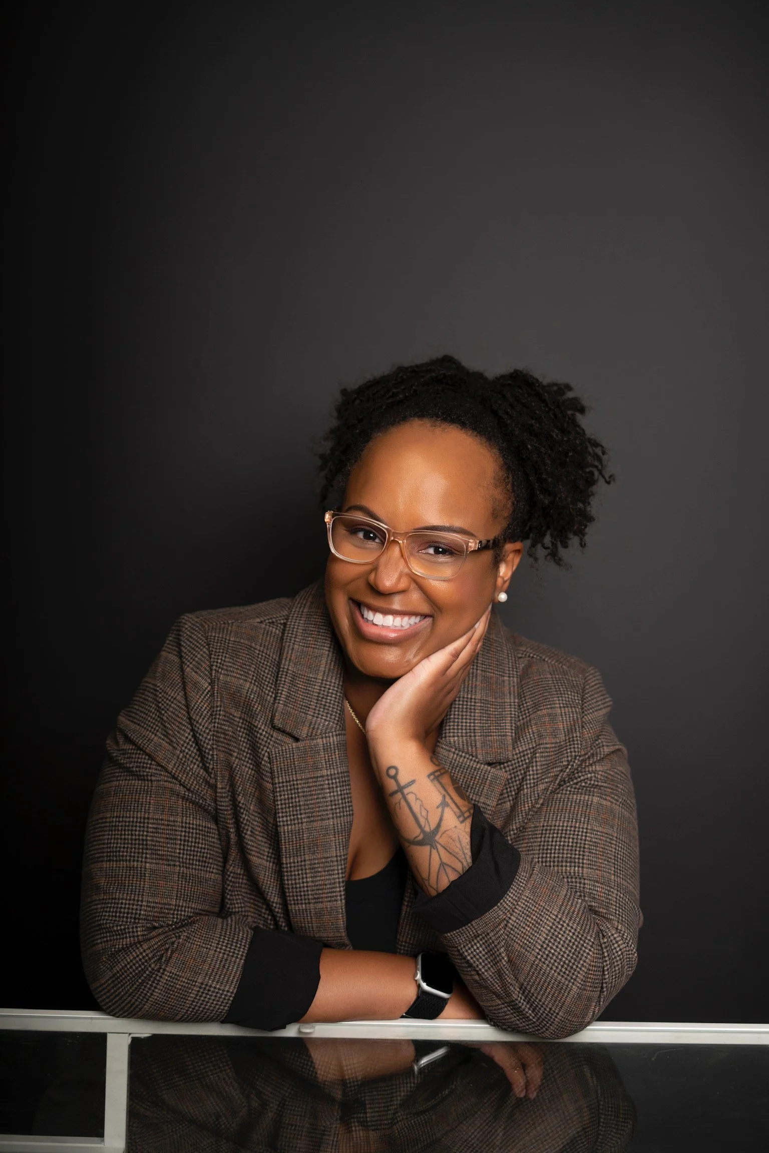 Portrait of a smiling woman with glasses, tattoos, and jewelry, resting her chin on her hand against a black background.